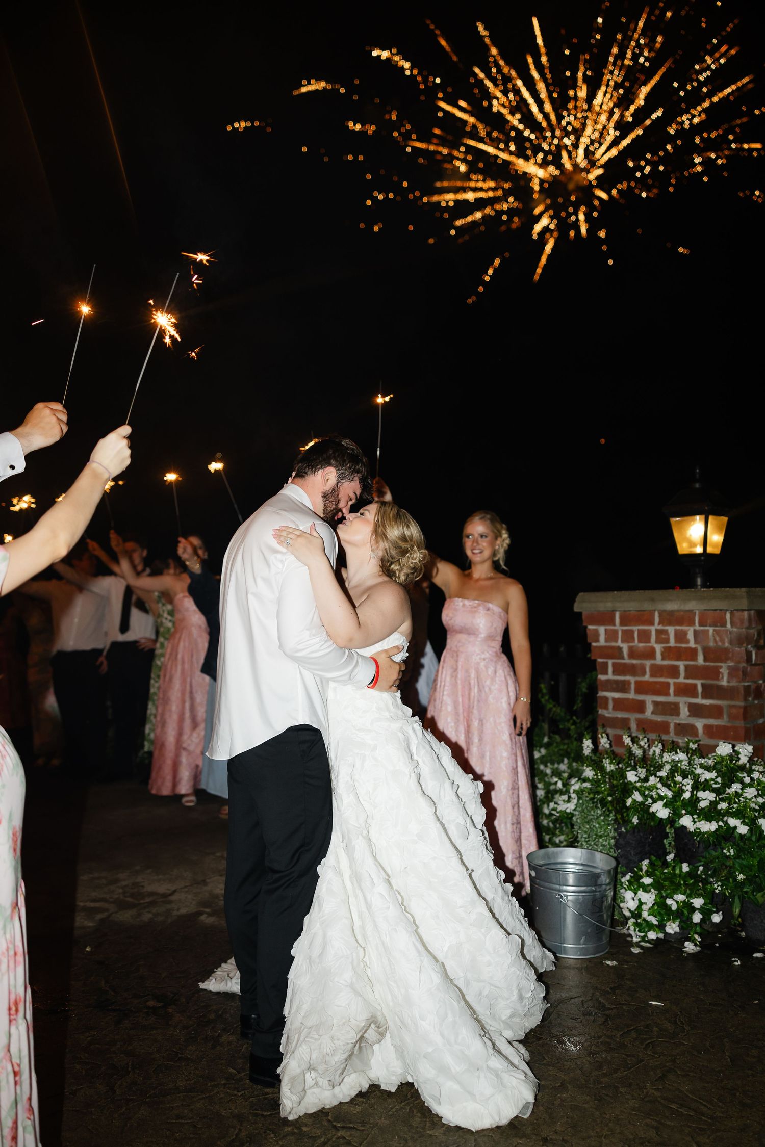 Bride and groom share a kiss during sparkler exit with fireworks lighting the night sky at outdoor wedding reception.