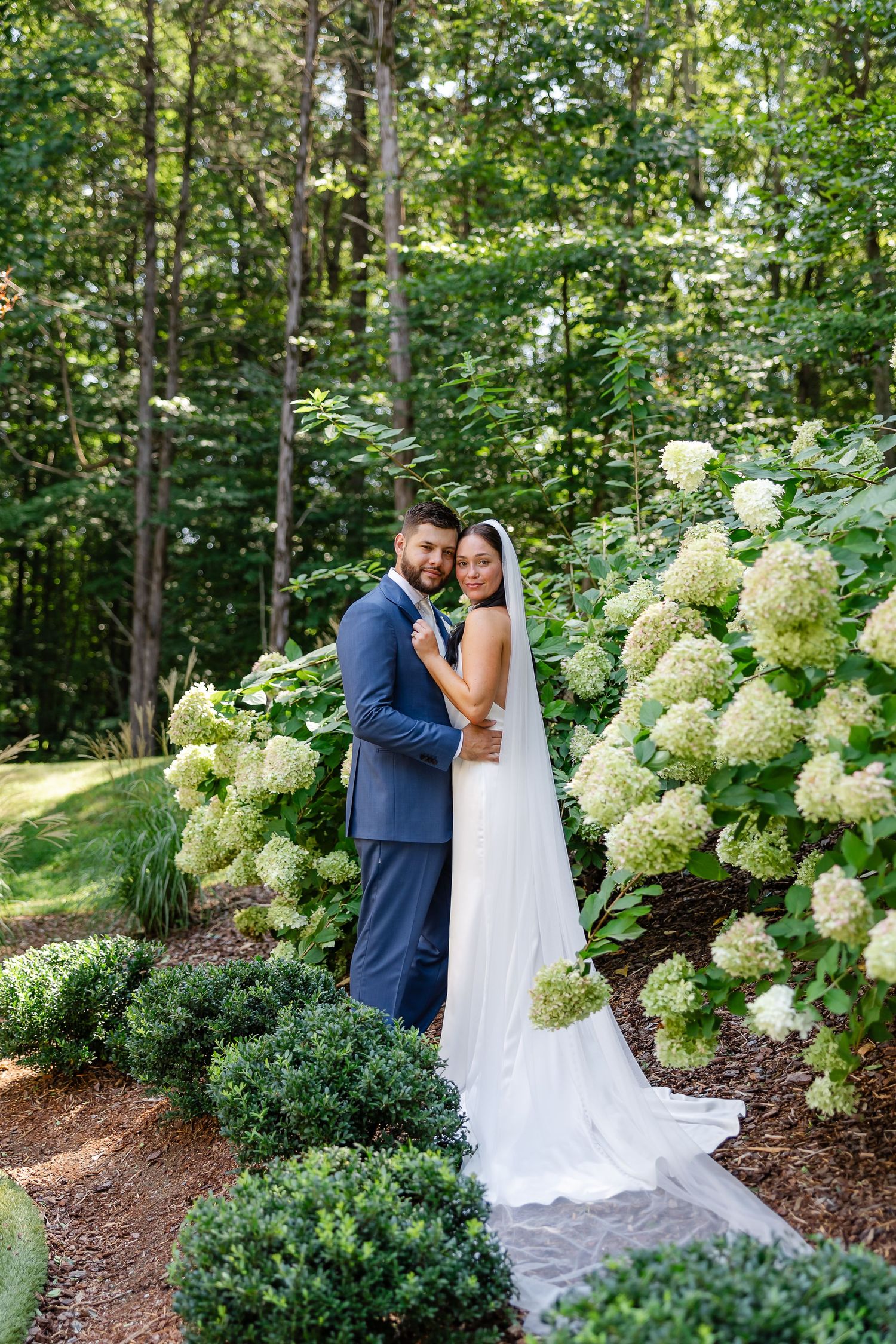 Bride and groom embracing in a lush garden surrounded by white hydrangeas and greenery on their wedding day. At FireFly Lane in Dickson, TN