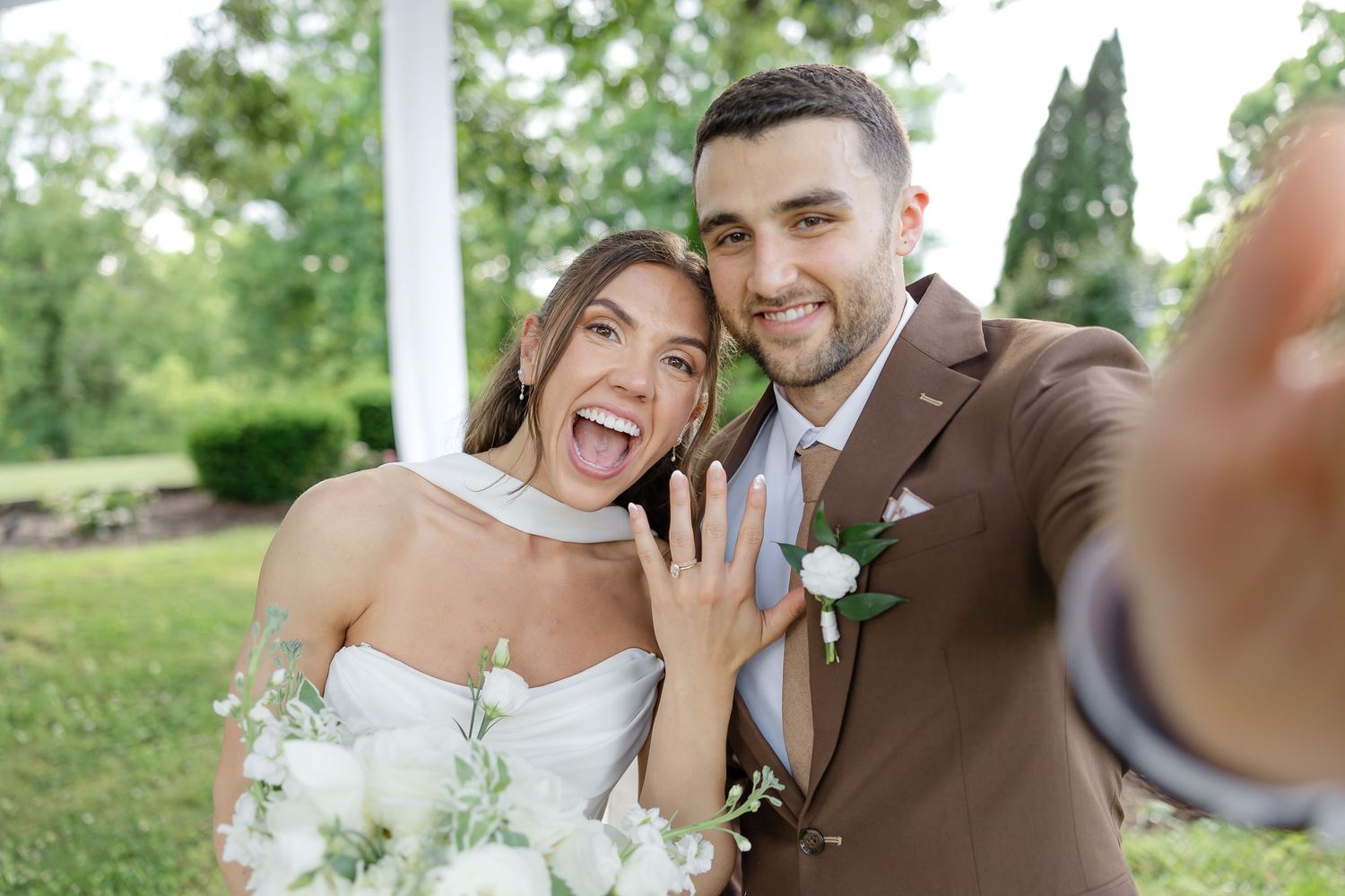 Happy newlyweds taking a selfie, bride showing wedding ring, holding white bouquet, groom in brown suit outdoors. at front porch farms in TN