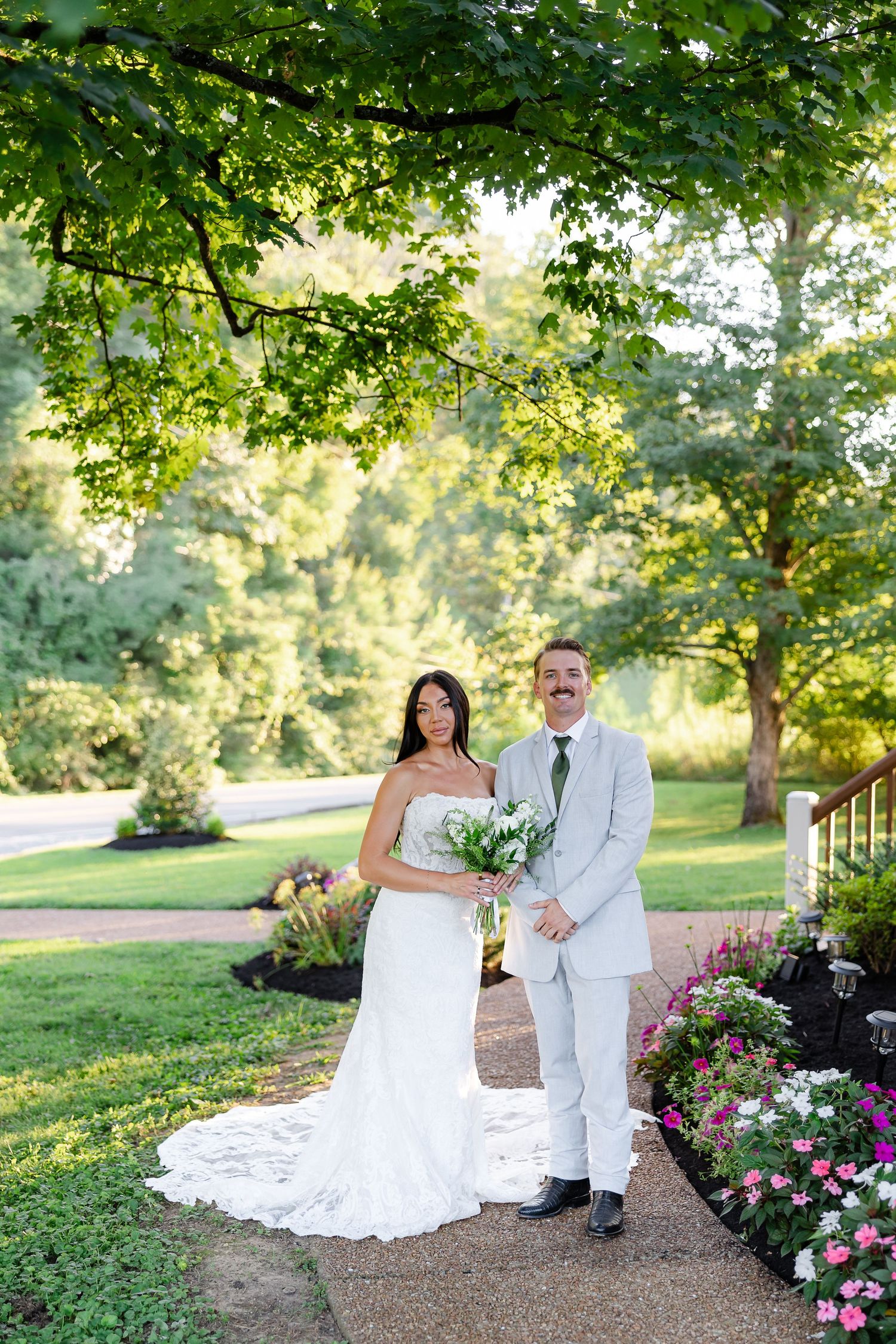 Bride and groom in gray suit pose together outdoors among colorful flowers and lush green trees.At Drakewood Farms in Nashville TN
