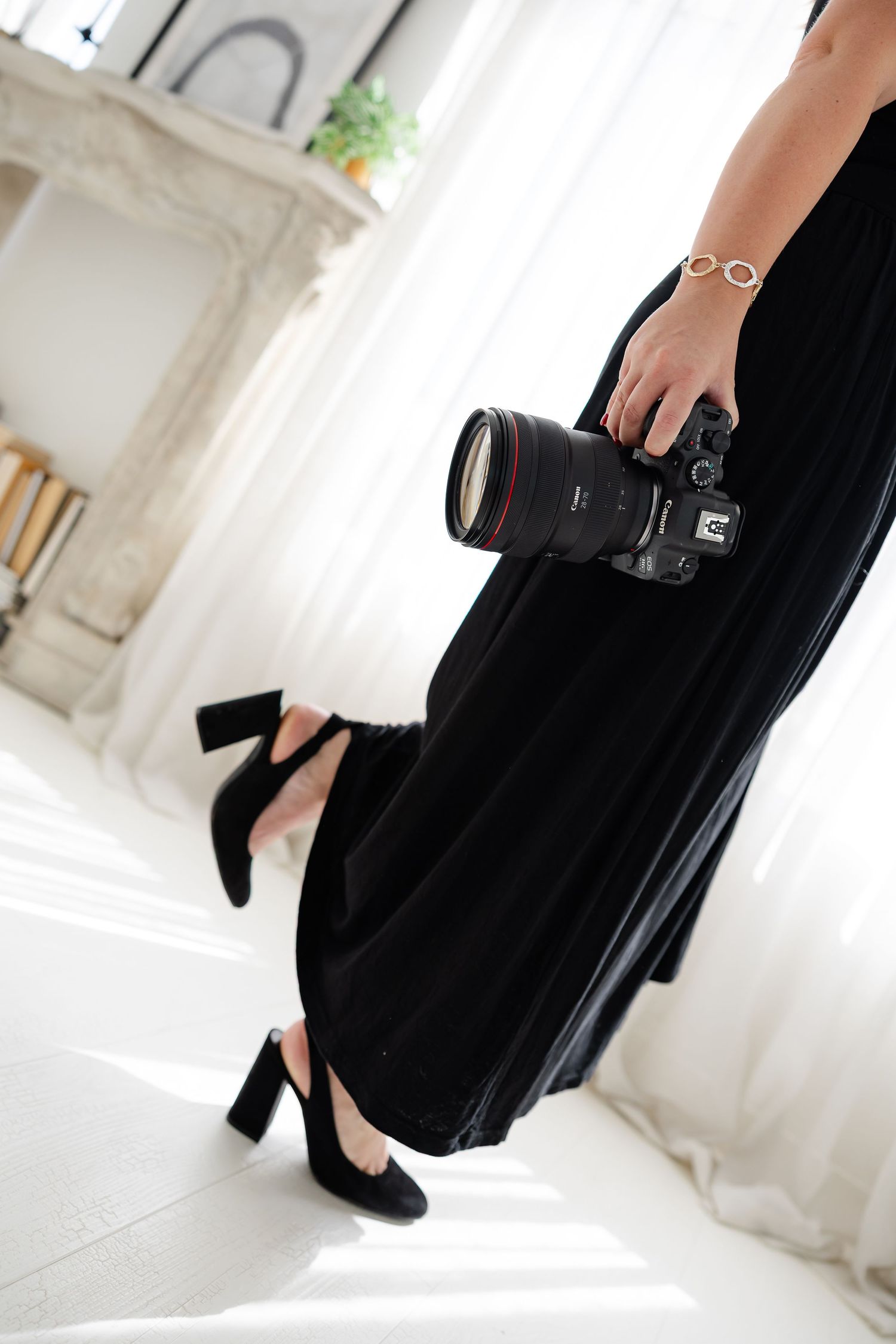 Woman in black dress and heels holding a Canon DSLR camera with zoom lens in a bright white studio.