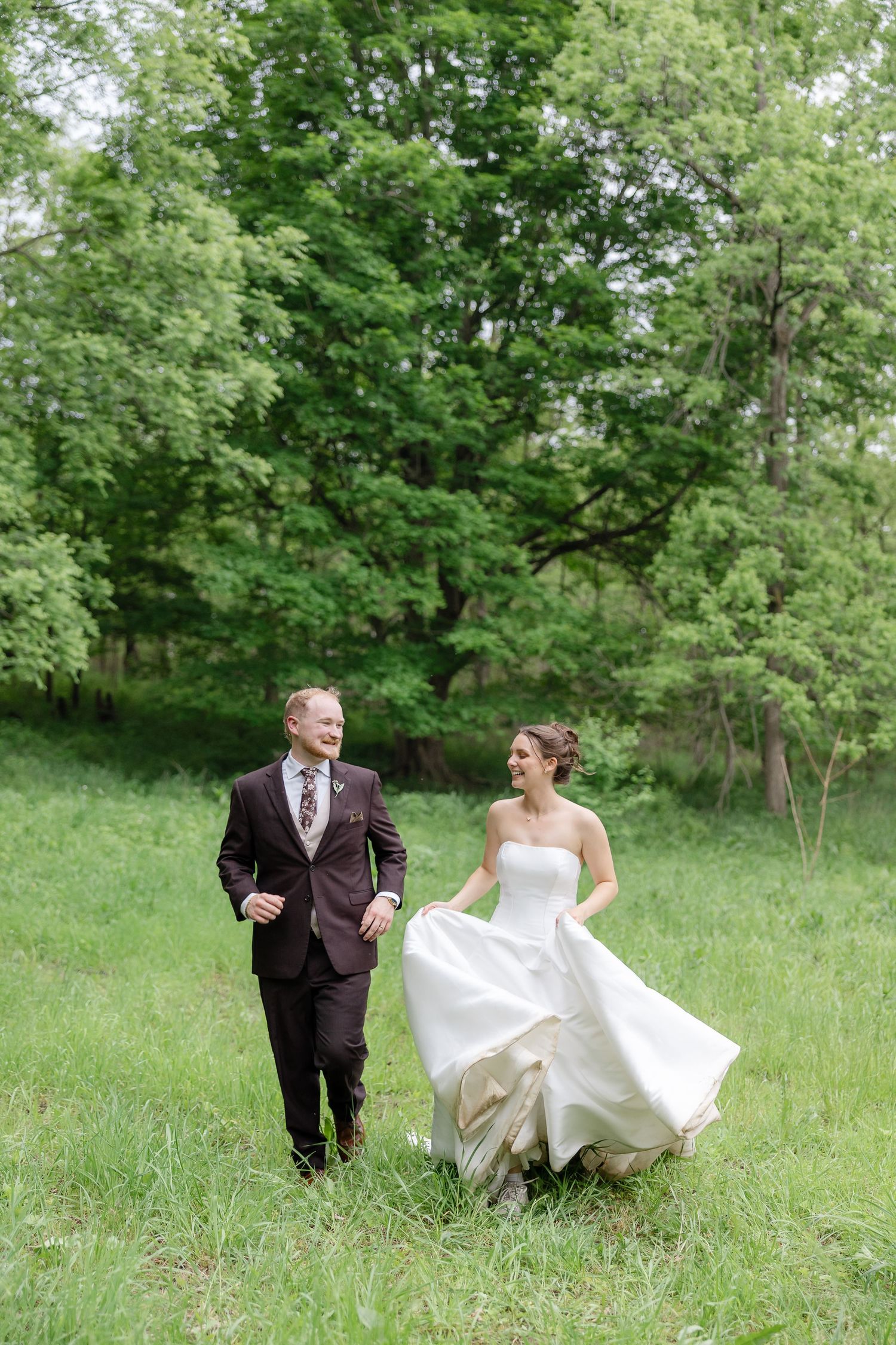 Bride and groom walking through lush green meadow, smiling joyfully, bride lifting white gown, groom in burgundy suit.