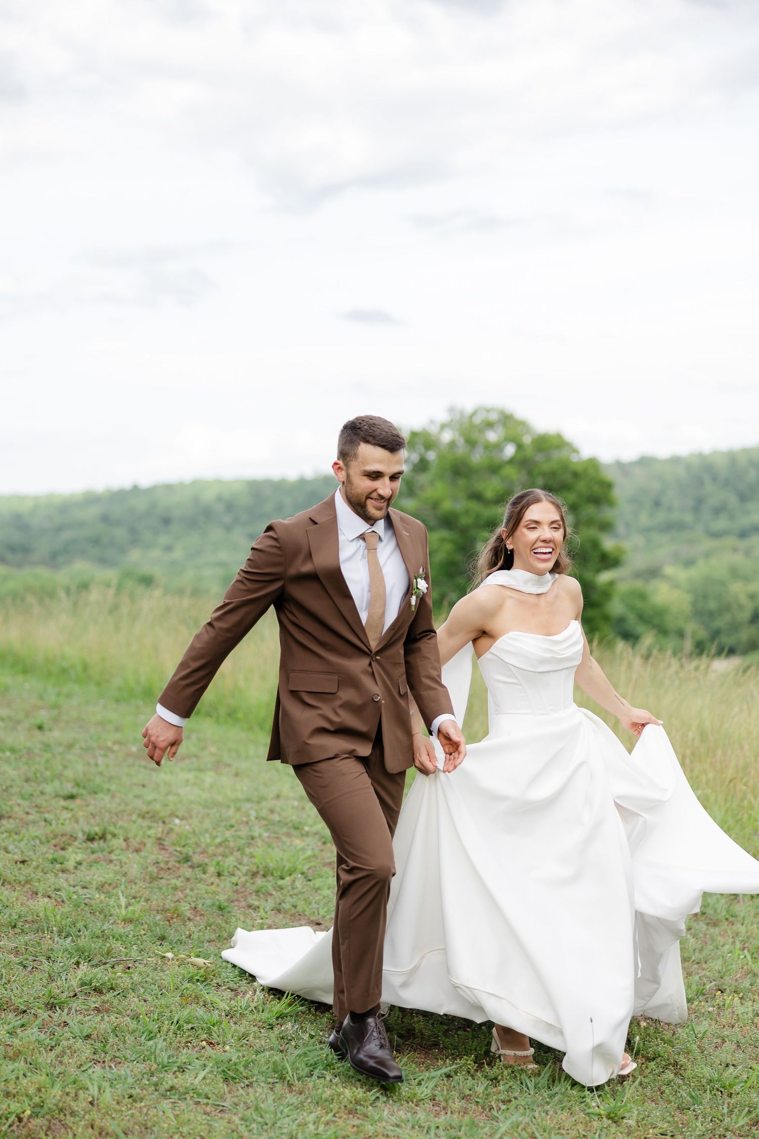 Smiling bride in white gown and groom in brown suit hold hands running through green field on their wedding day.