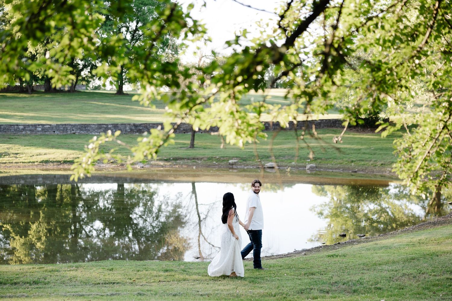 Bride and groom holding hands walking by a serene pond surrounded by lush green trees on their wedding day.