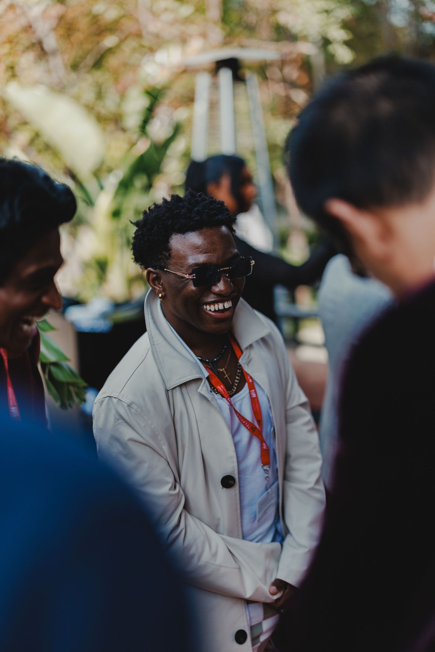 Smiling man in sunglasses and white jacket wearing a lanyard at an outdoor social gathering.