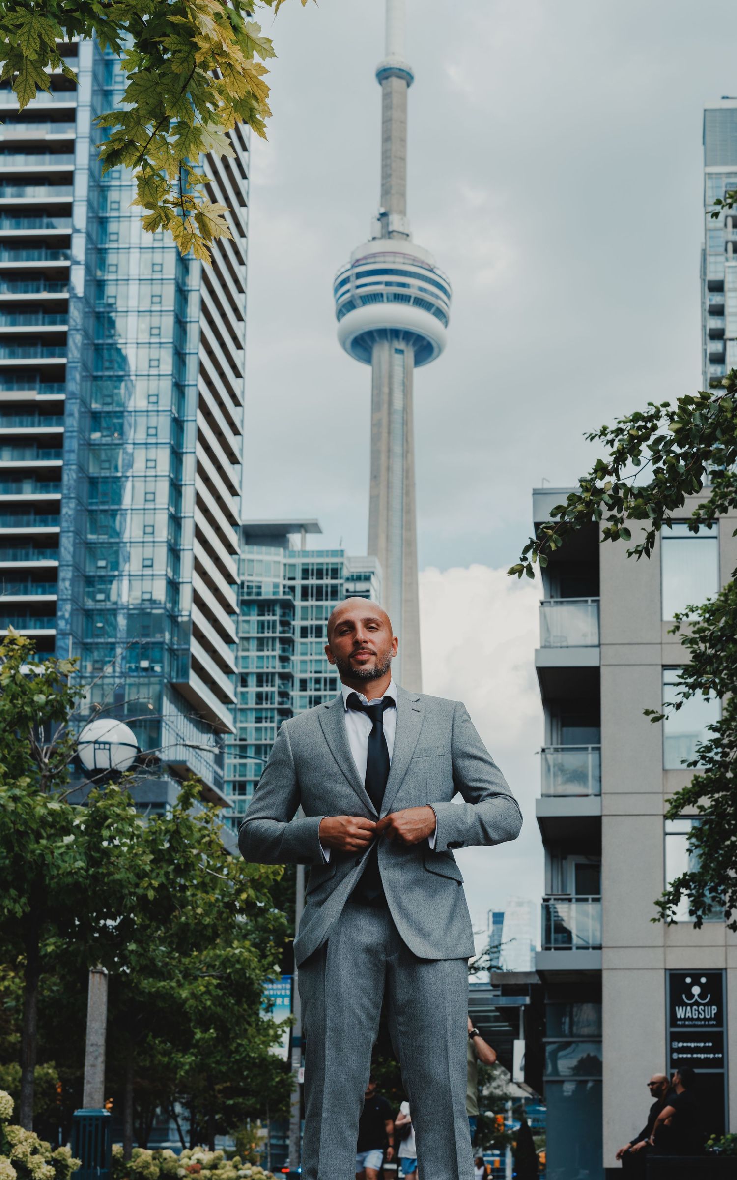 Man in gray suit standing before CN Tower and Toronto skyline on a cloudy day.