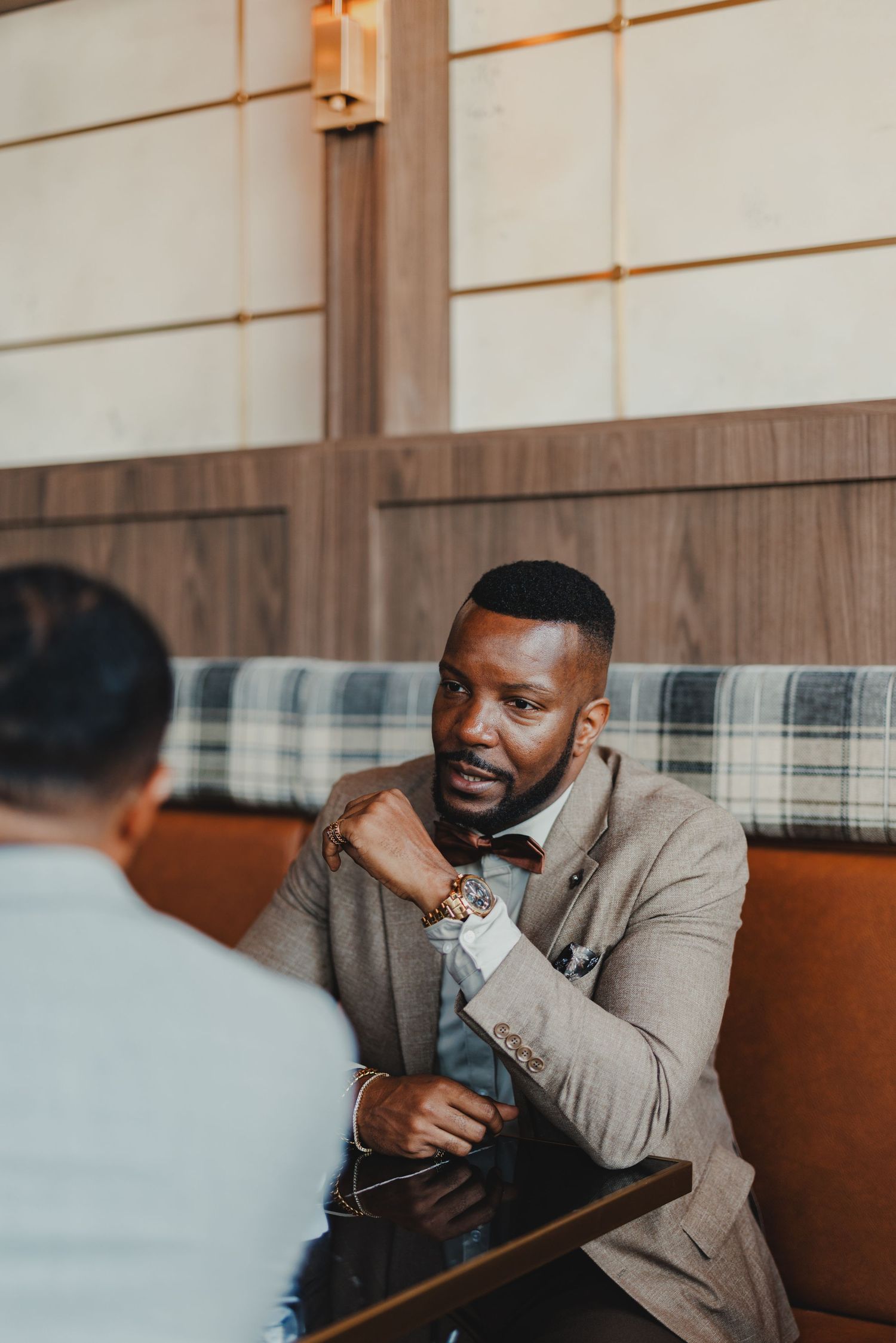 Well-dressed Black man in gray suit and bow tie sits in restaurant booth, engaged in conversation with someone across the table.