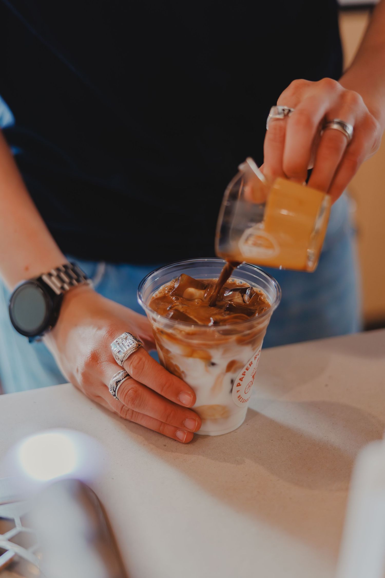 Hands pouring espresso shot into an iced latte in a clear plastic cup on a café counter.