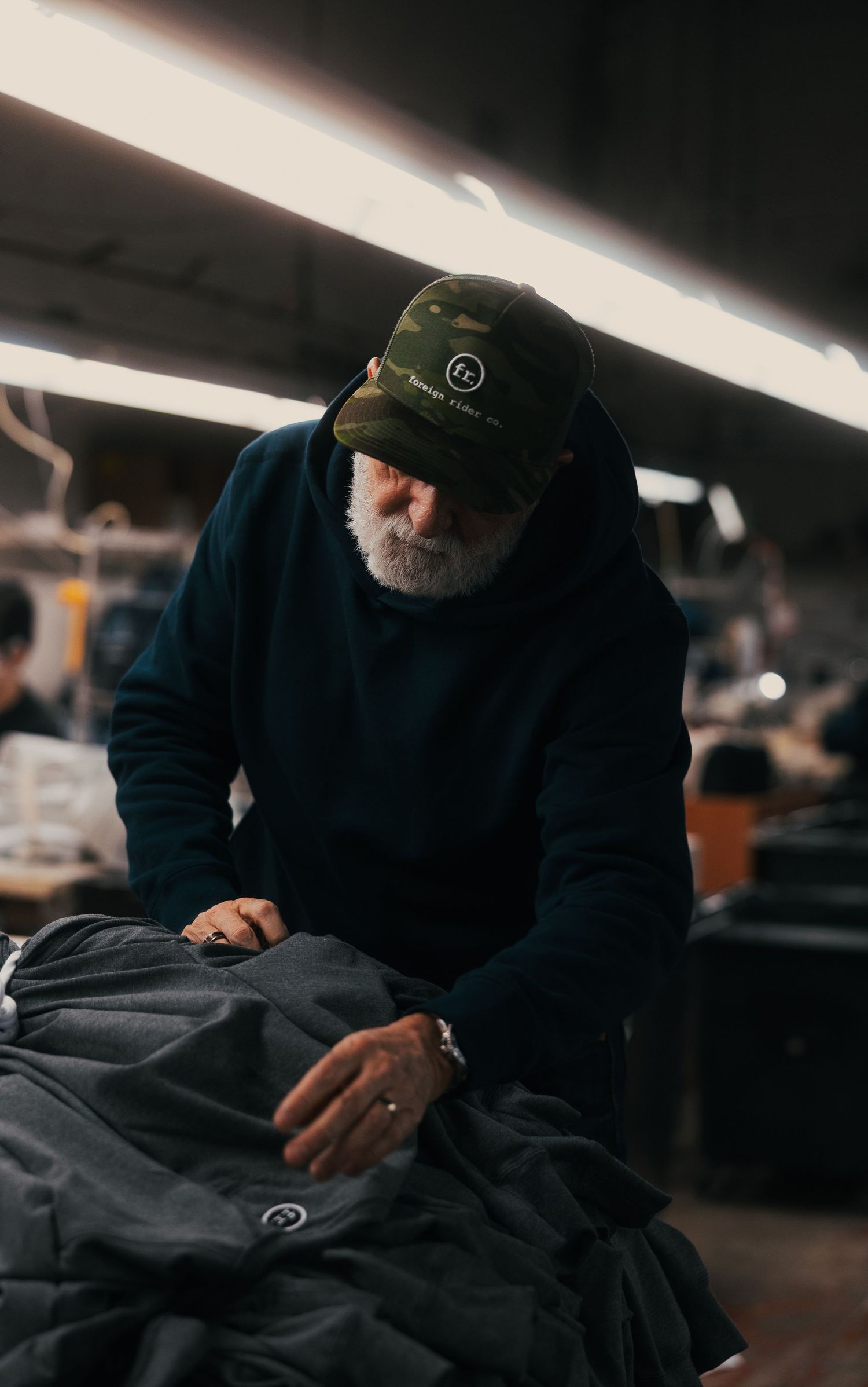 Elderly bearded man in camo cap inspecting dark fabric garments in a professional textile manufacturing workshop.