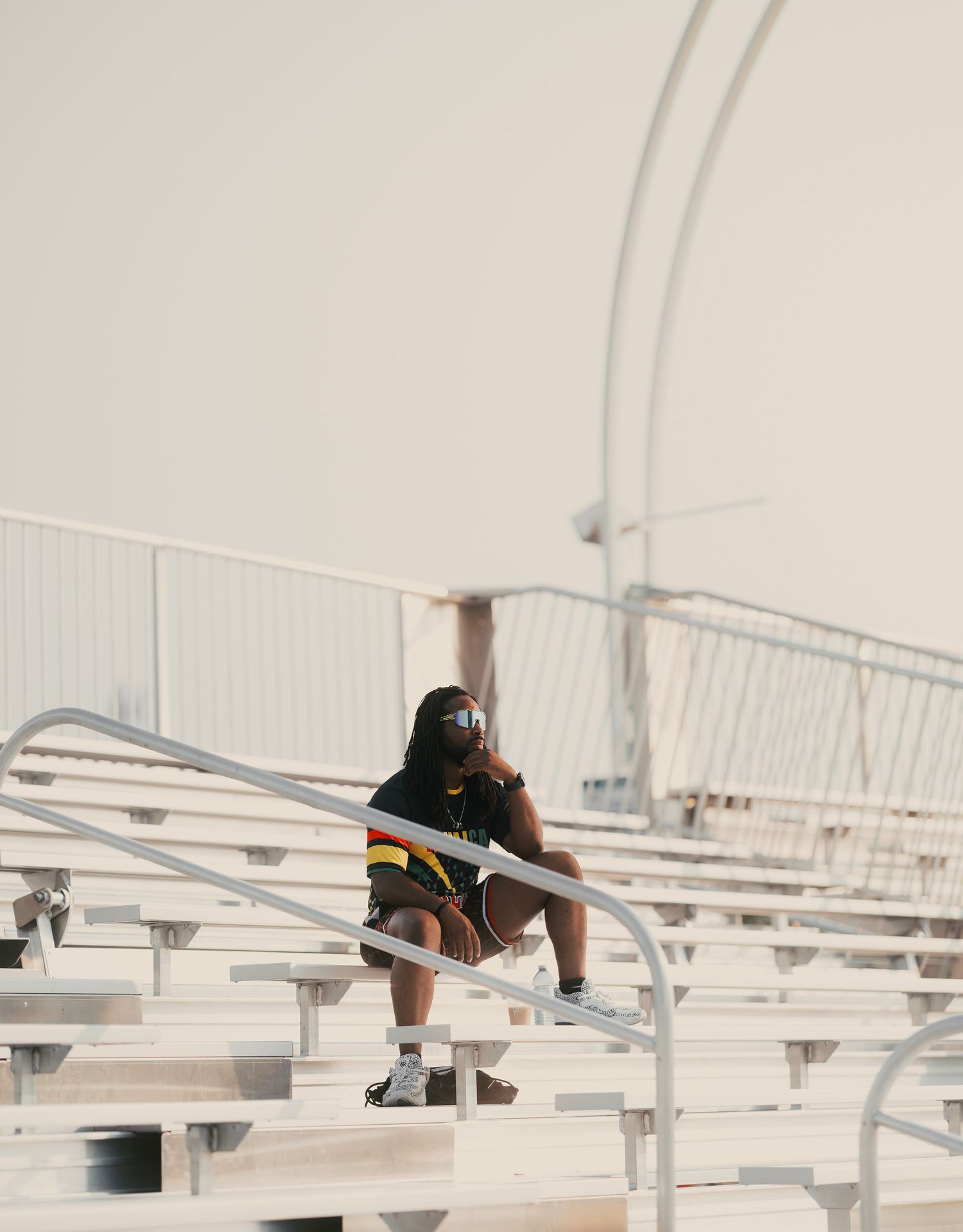 Person sitting alone on metal bleachers outdoors, wearing casual athletic wear and sunglasses on a hazy day.