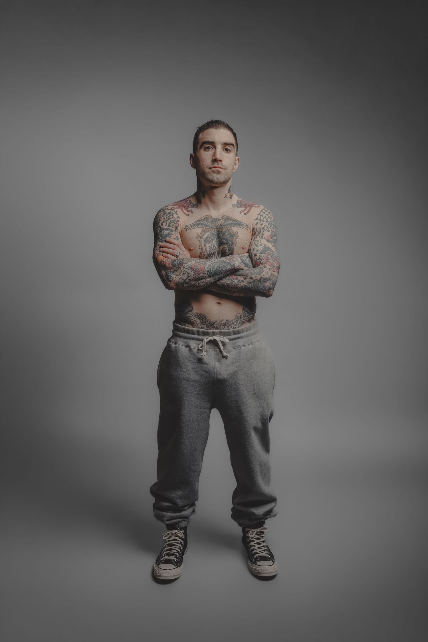 Tattooed shirtless man standing with arms crossed wearing grey sweatpants and black Converse sneakers against grey background.