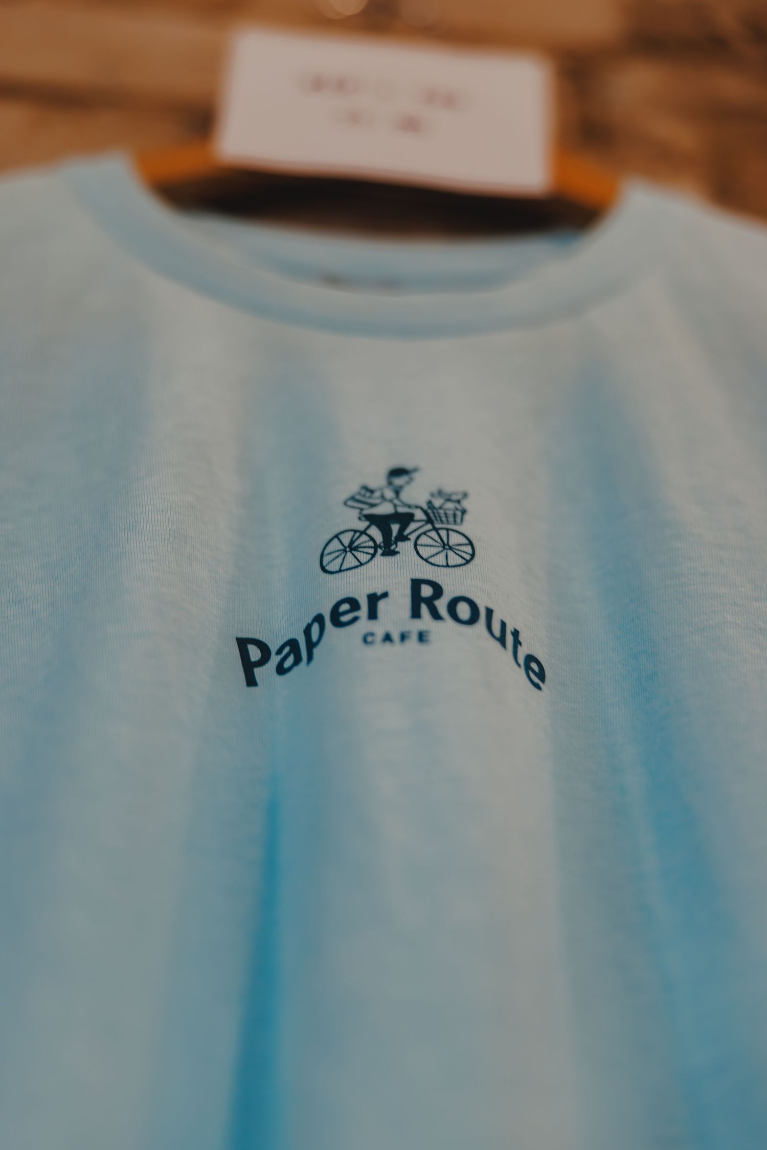 Light blue Paper Route Cafe branded t-shirt with bicycle logo hanging on a wooden hanger.