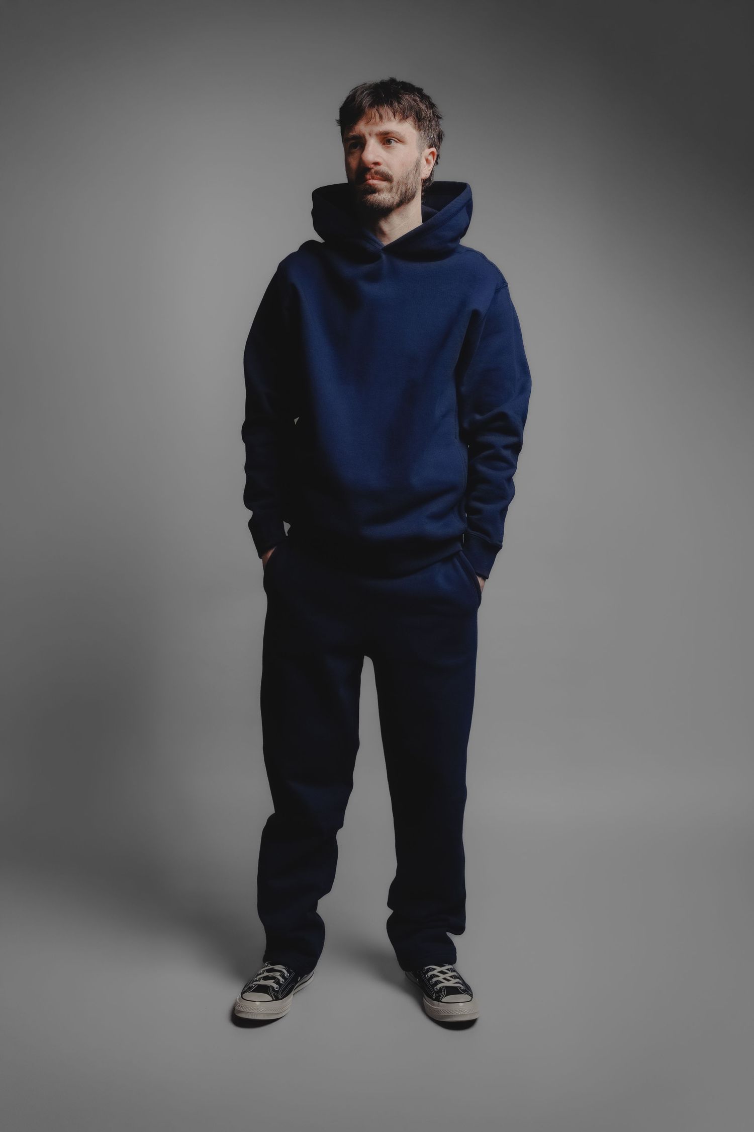 Man wearing navy blue hoodie and dark joggers with sneakers, standing against gray background.