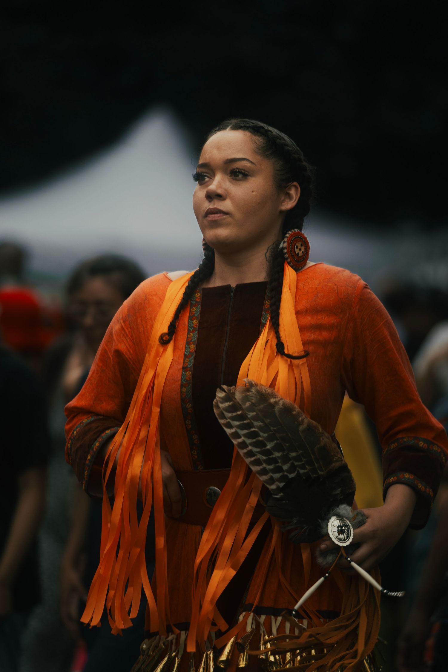 Woman in orange fringed Native American regalia with braids and feathered fan at outdoor cultural event.