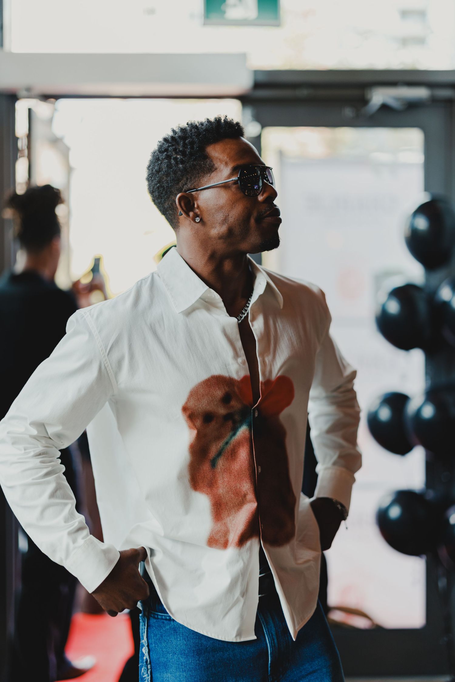 Stylish man in white shirt with dog print, sunglasses, and jeans posing confidently indoors near black balloons.