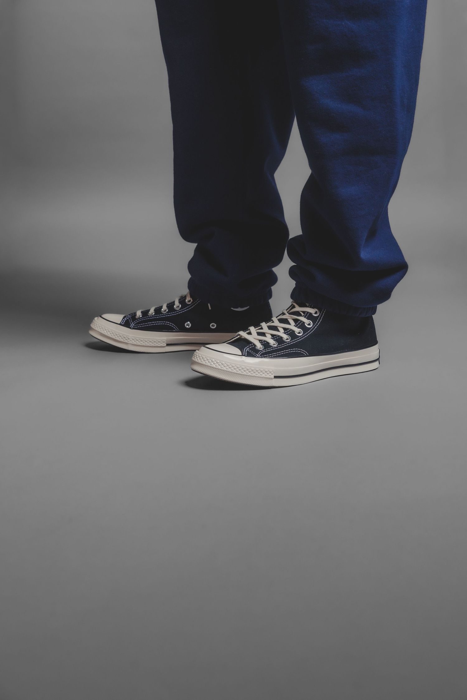 Close-up of feet wearing black Converse Chuck Taylor 70 sneakers with blue and black pants on grey floor.