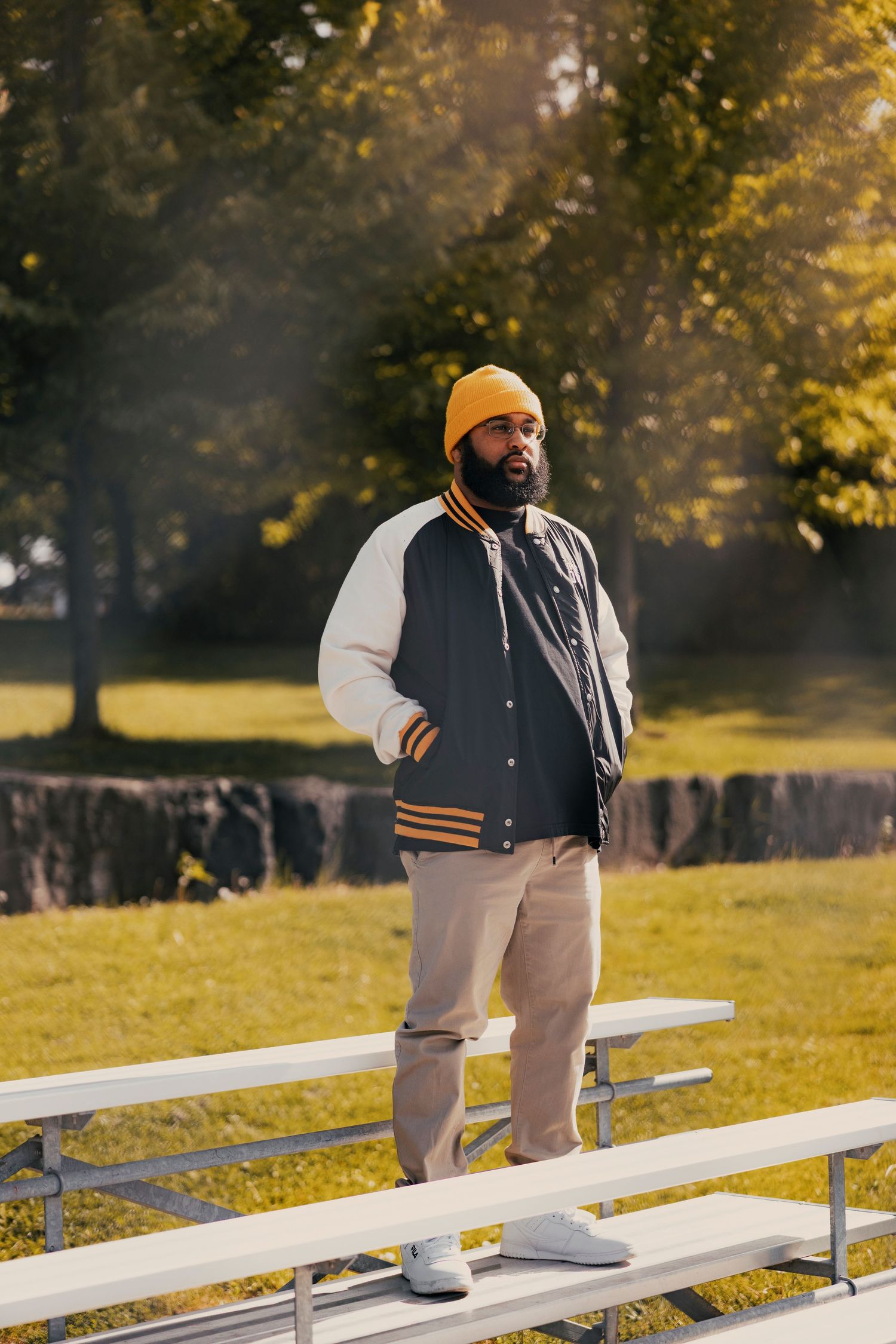 Man in yellow beanie and varsity jacket standing on bleachers in a sunny park.