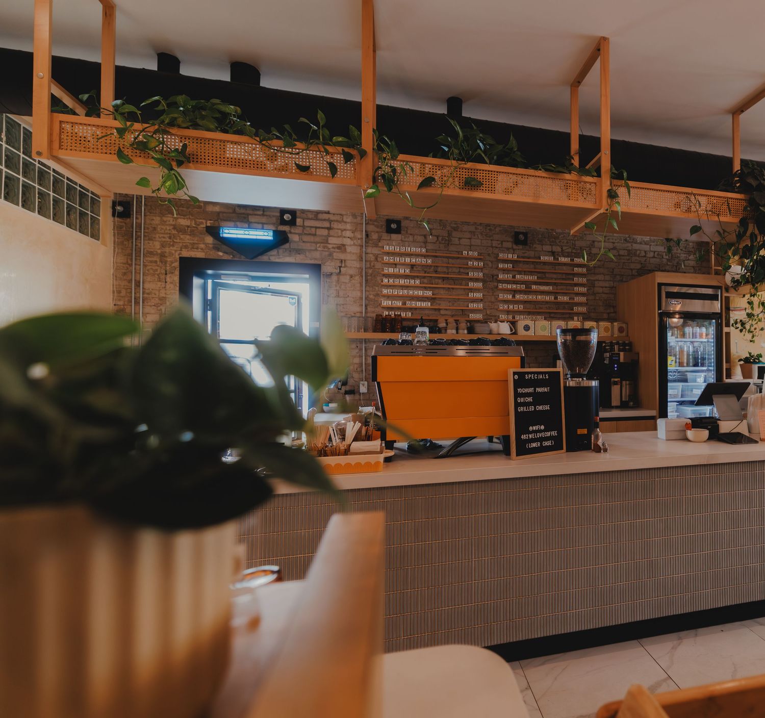 Cozy coffee shop interior with exposed brick wall, hanging plants, wooden shelving, and an espresso bar with menu boards.