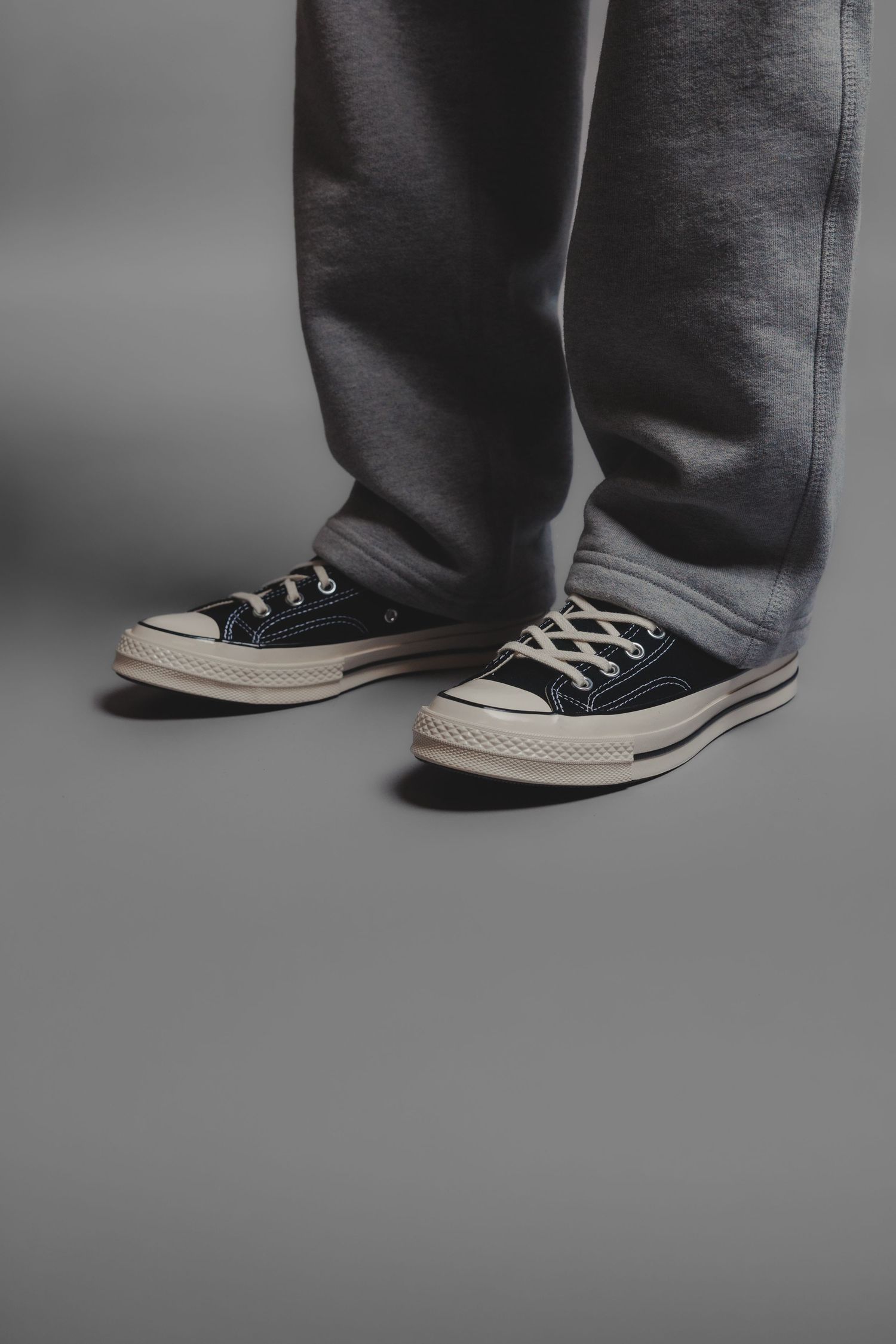 Close-up of grey sweatpants and black Converse Chuck Taylor low-top sneakers on grey background.