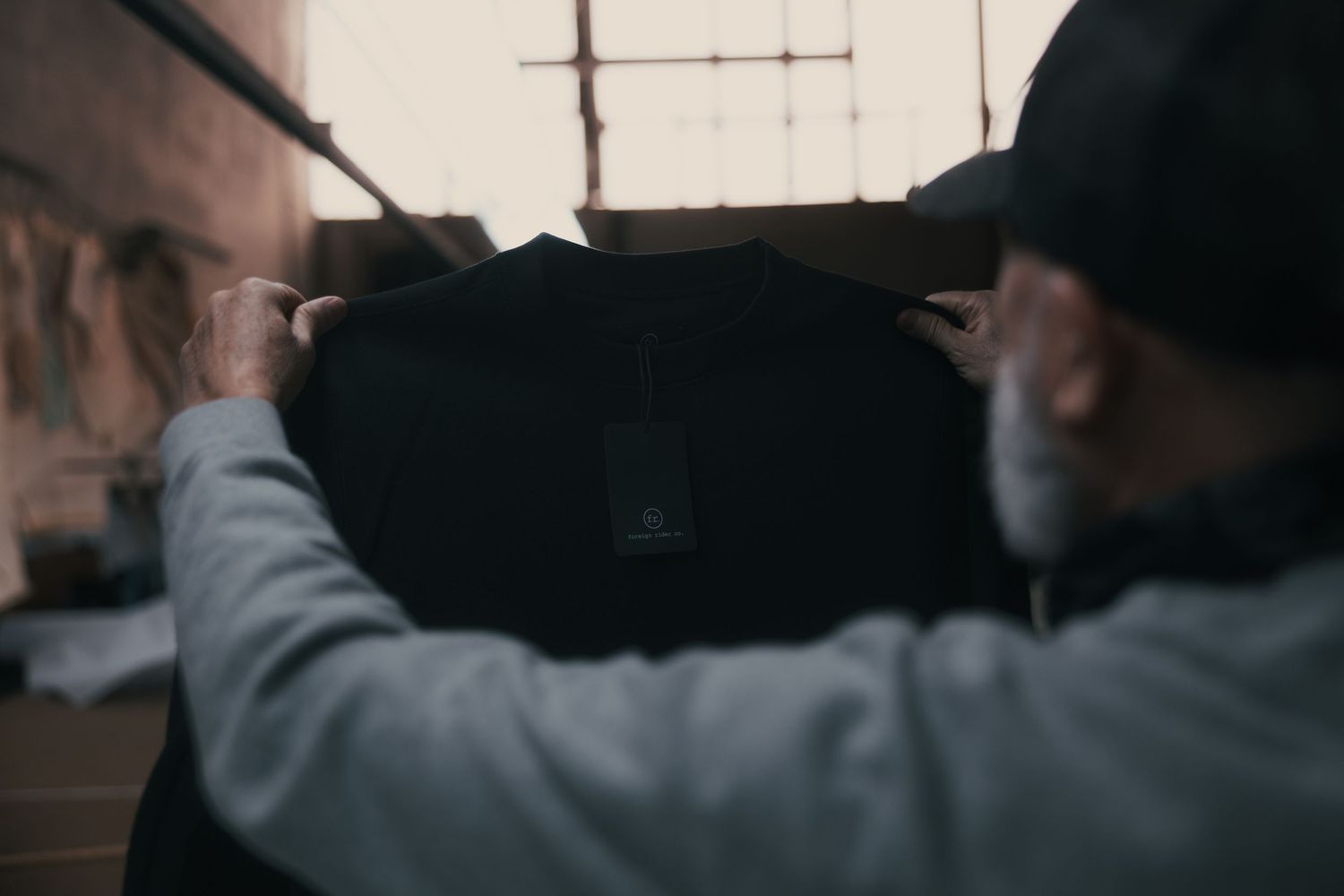 Man in gray hoodie and black cap holding a dark jacket in a dimly lit industrial space.