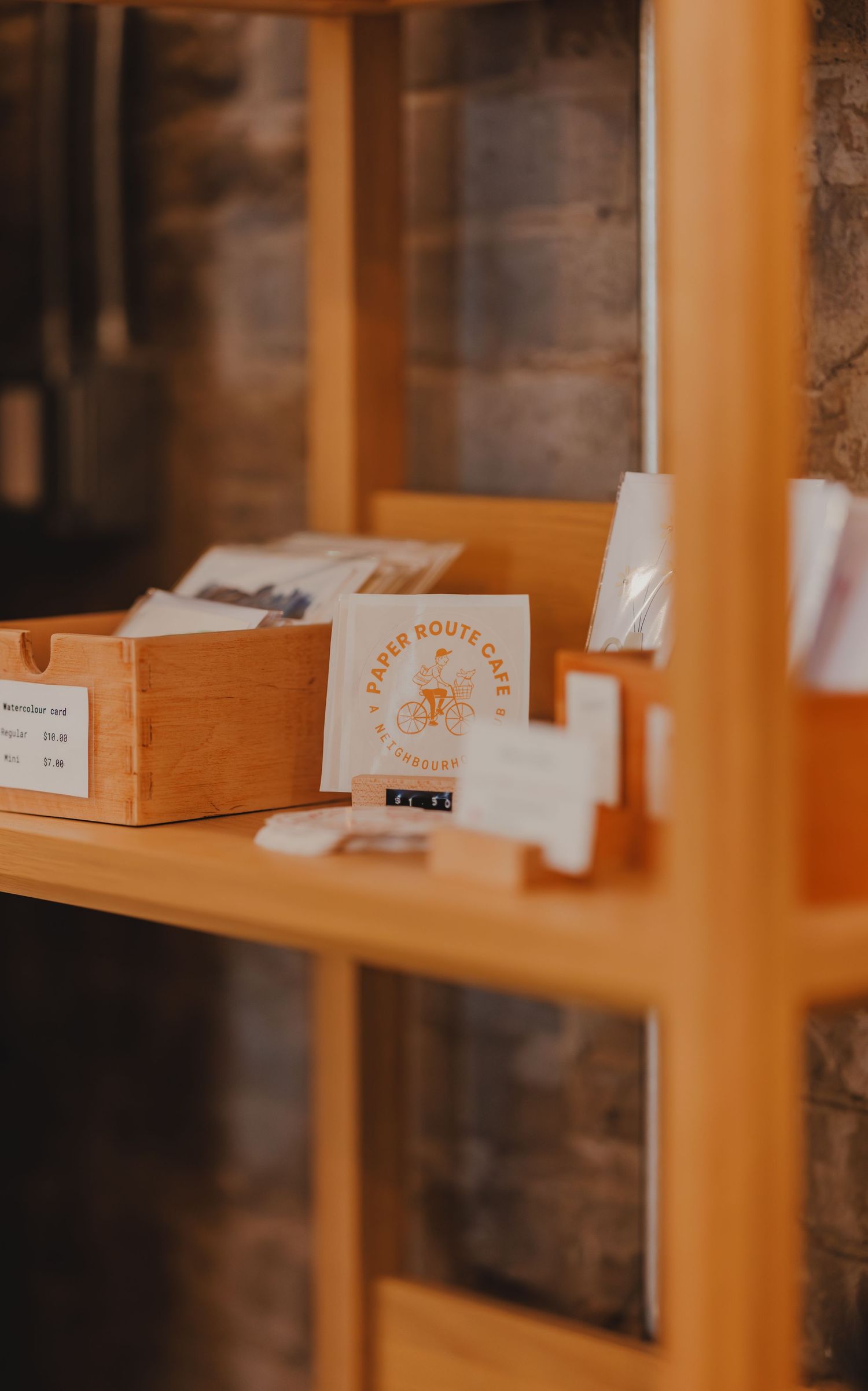 Wooden retail shelf displaying Paper Route Cafe branded merchandise, cards, and packaged goods.