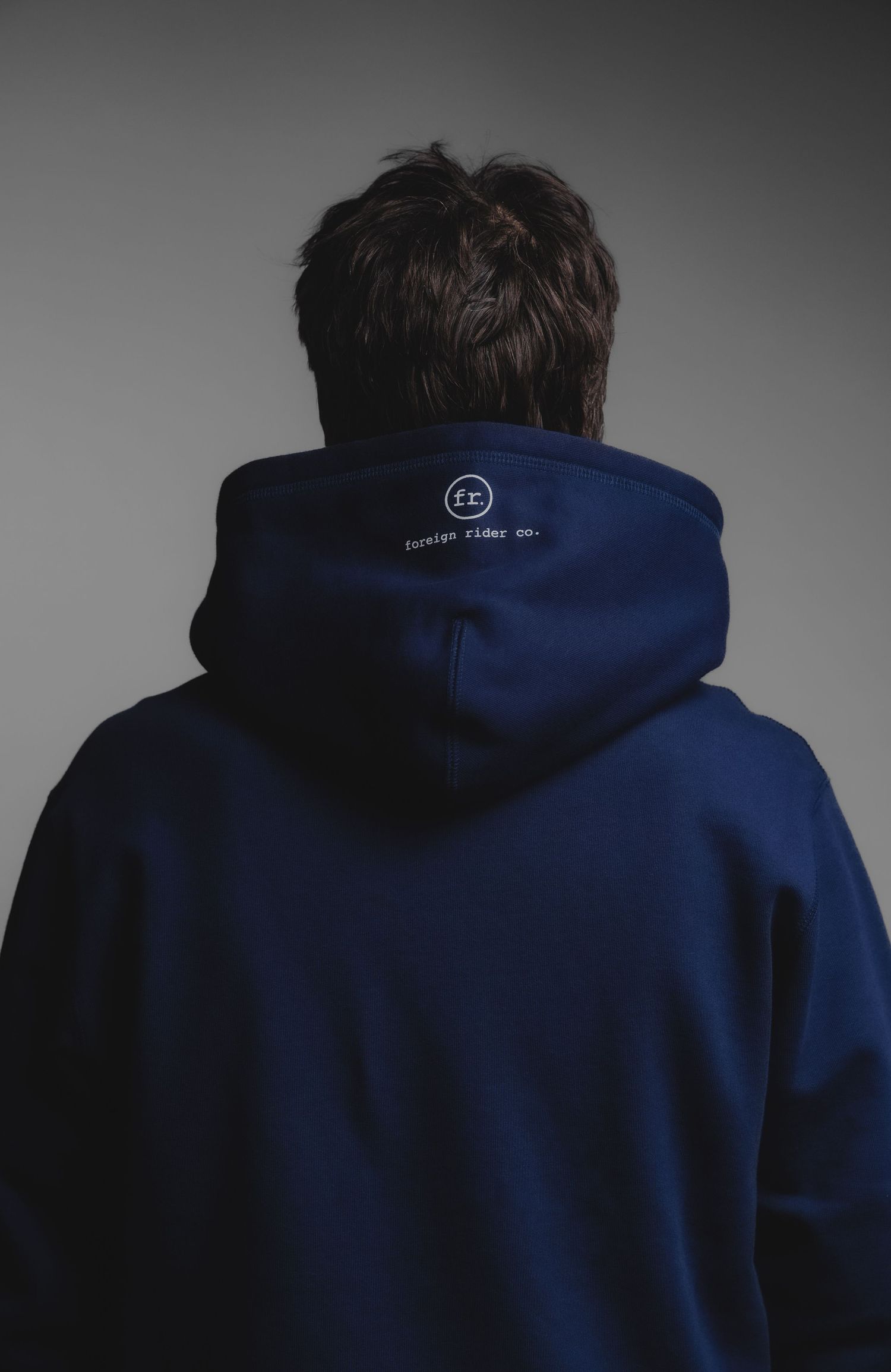 Back view of a man wearing a navy blue hoodie with a small logo on the hood, against a gray background.