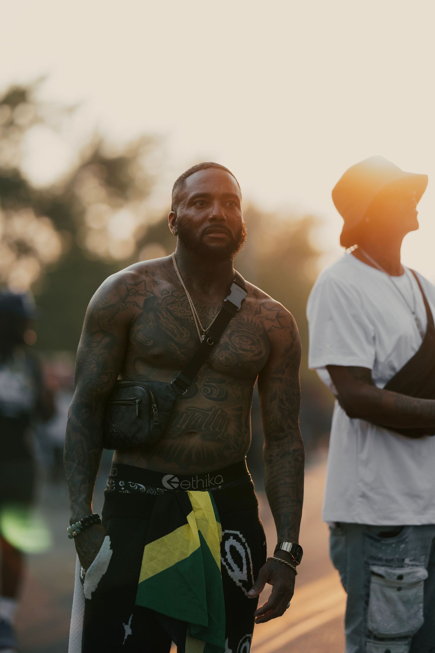 Heavily tattooed shirtless man with fanny pack stands outdoors at golden hour with crowd in background.