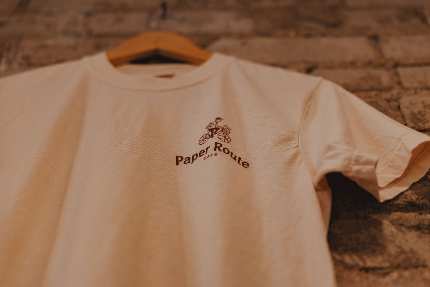 White Paper Route cafe branded t-shirt on a wooden hanger against a rustic brick wall background.