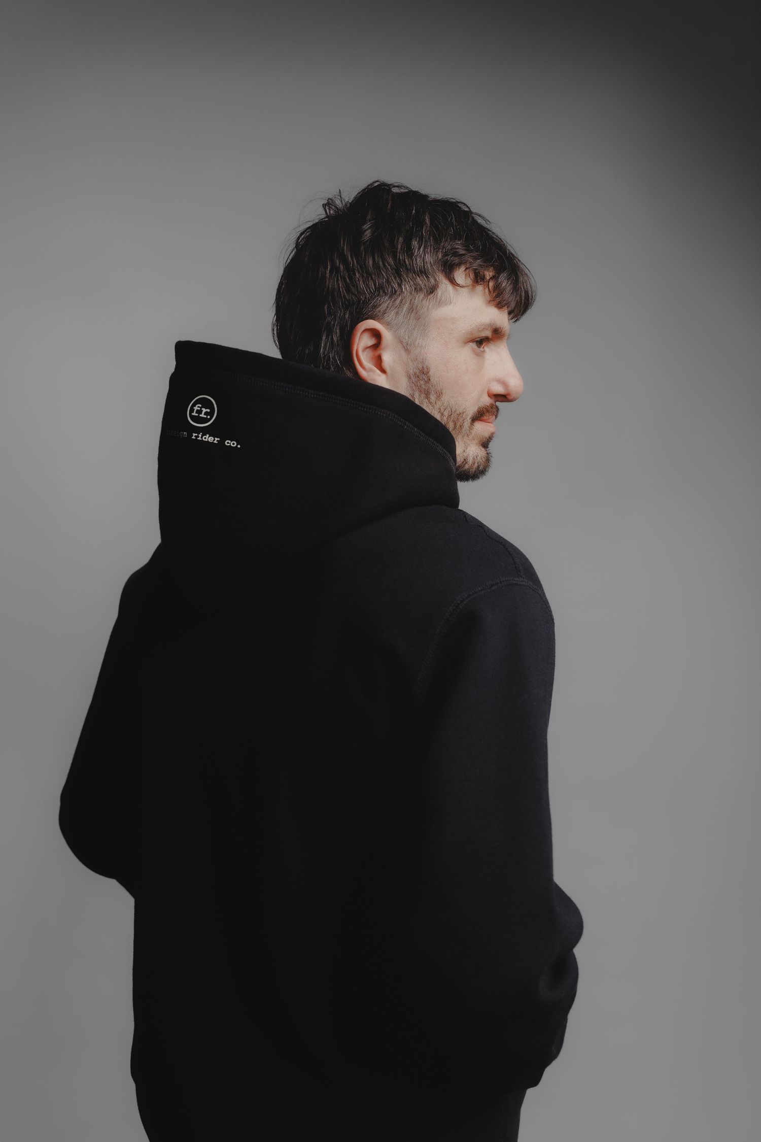 Man in black hoodie with logo detail, shot from behind against gray background.