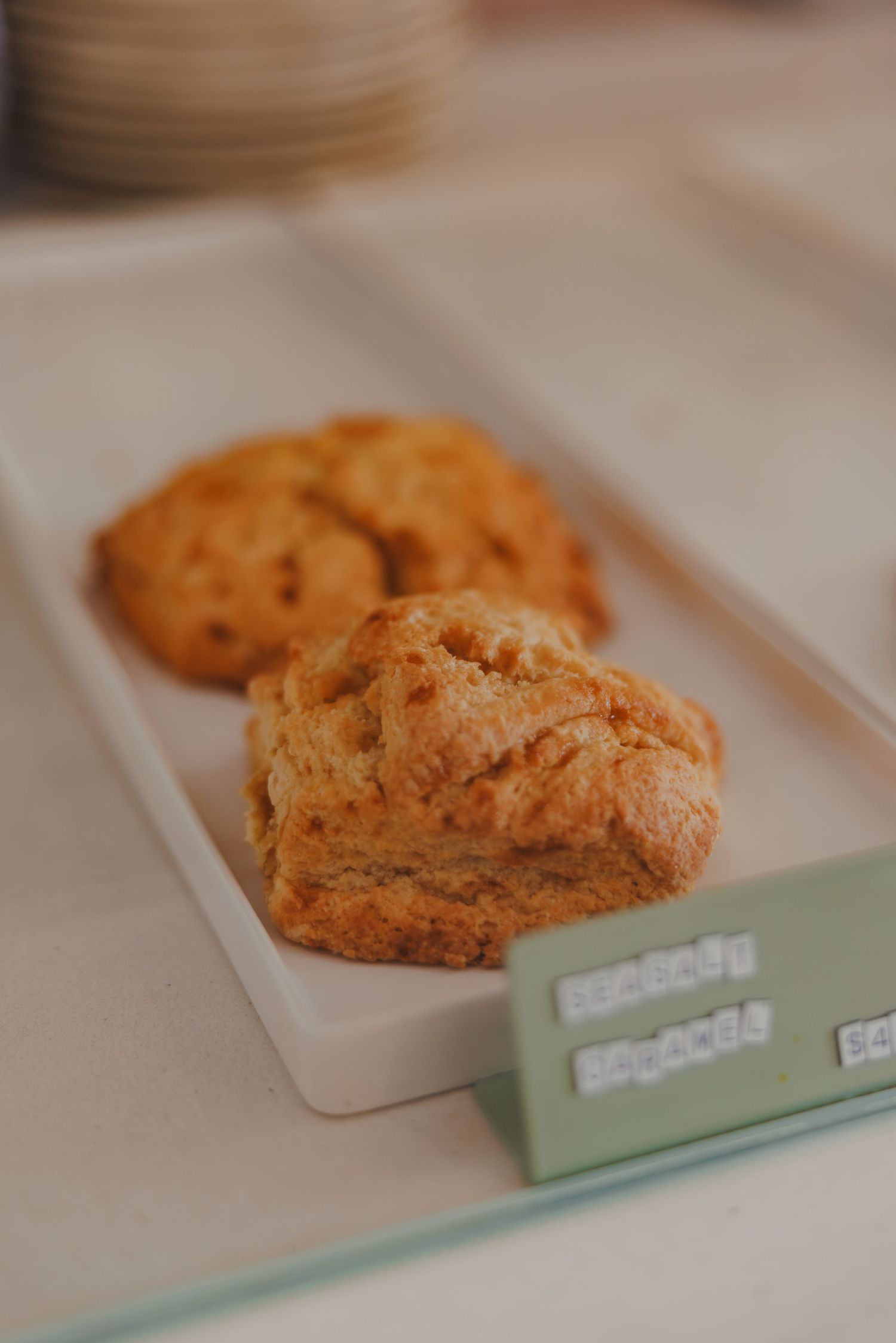 Two golden-brown scones on a white rectangular tray with a small label sign on a cafe counter.