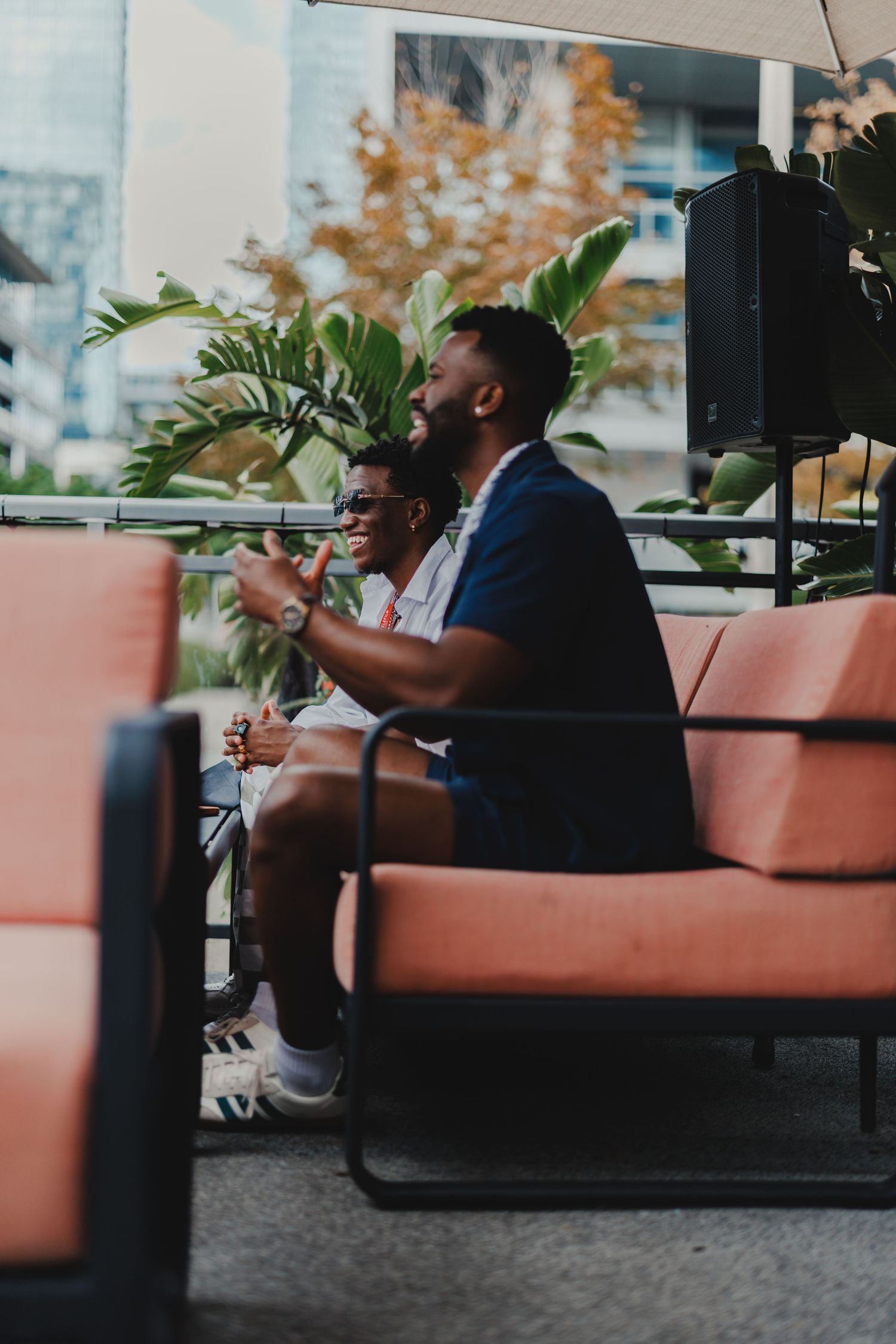 Two men sitting on orange outdoor furniture in an urban setting, enjoying a relaxed conversation surrounded by greenery.