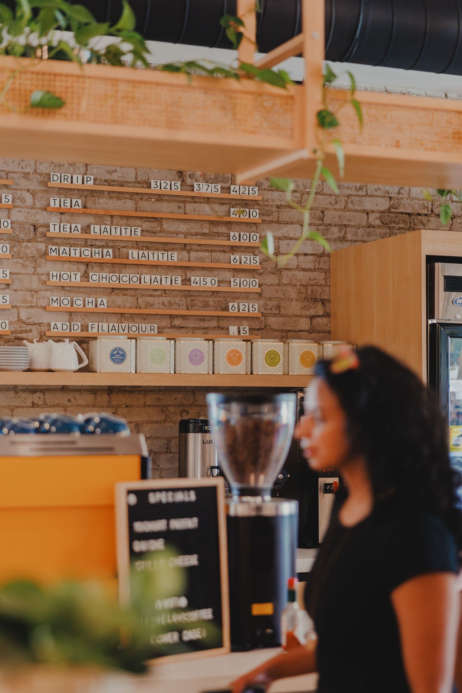 Barista working at a modern cafe counter with brick wall menu board and coffee grinder in background.