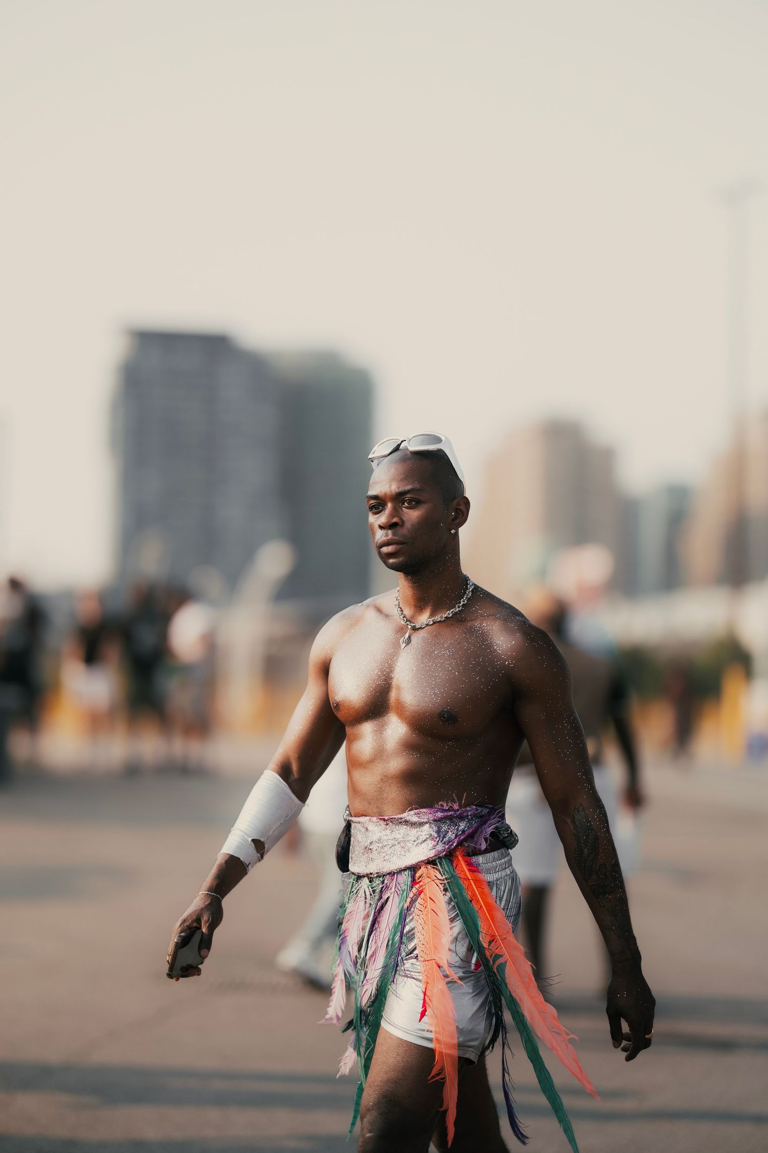 Shirtless athletic Black man in colorful costume walking at outdoor Pride event with city skyline.