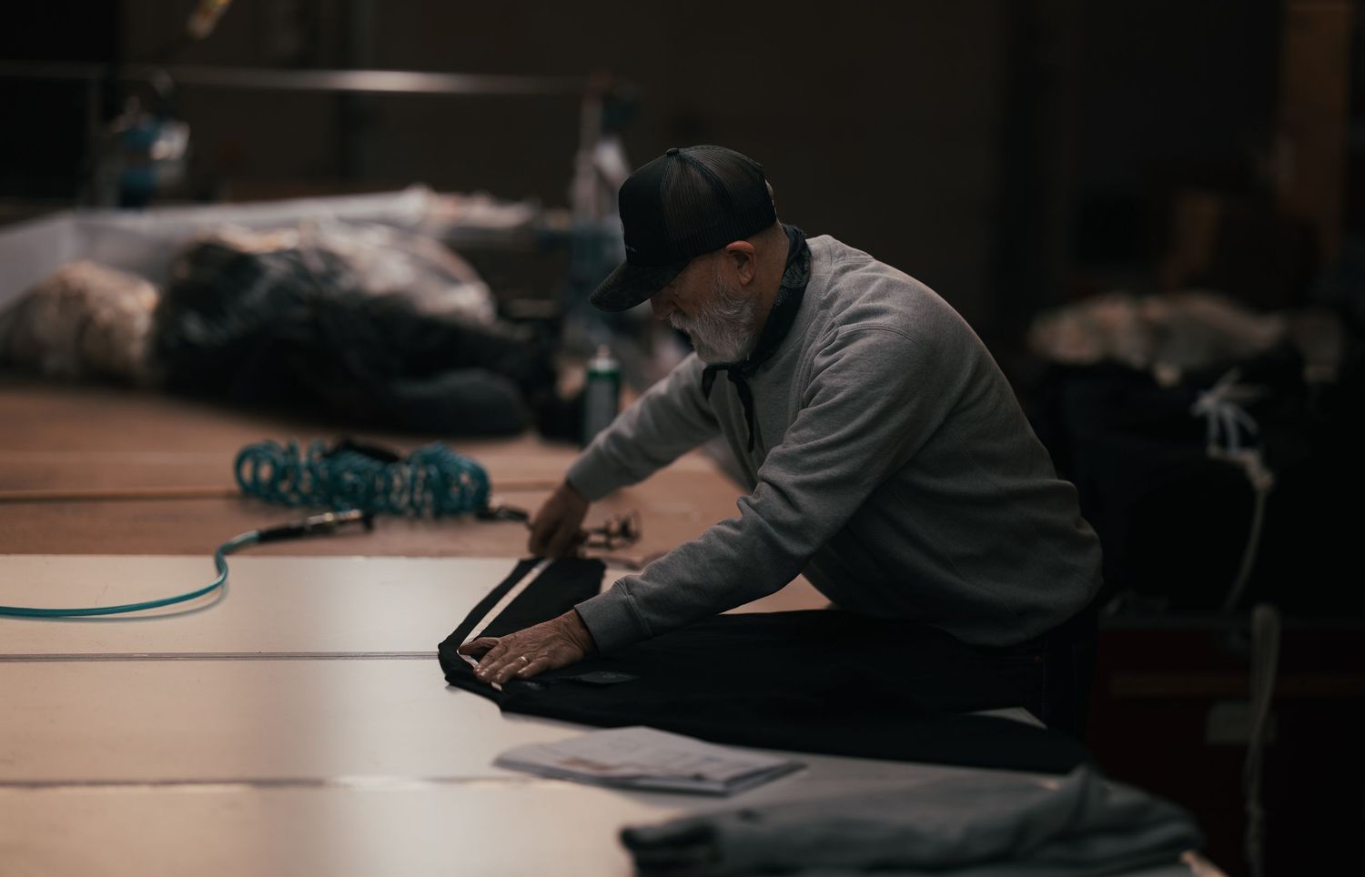 Elderly man in cap sits on wooden floor working with dark fabric in a dimly lit workshop.
