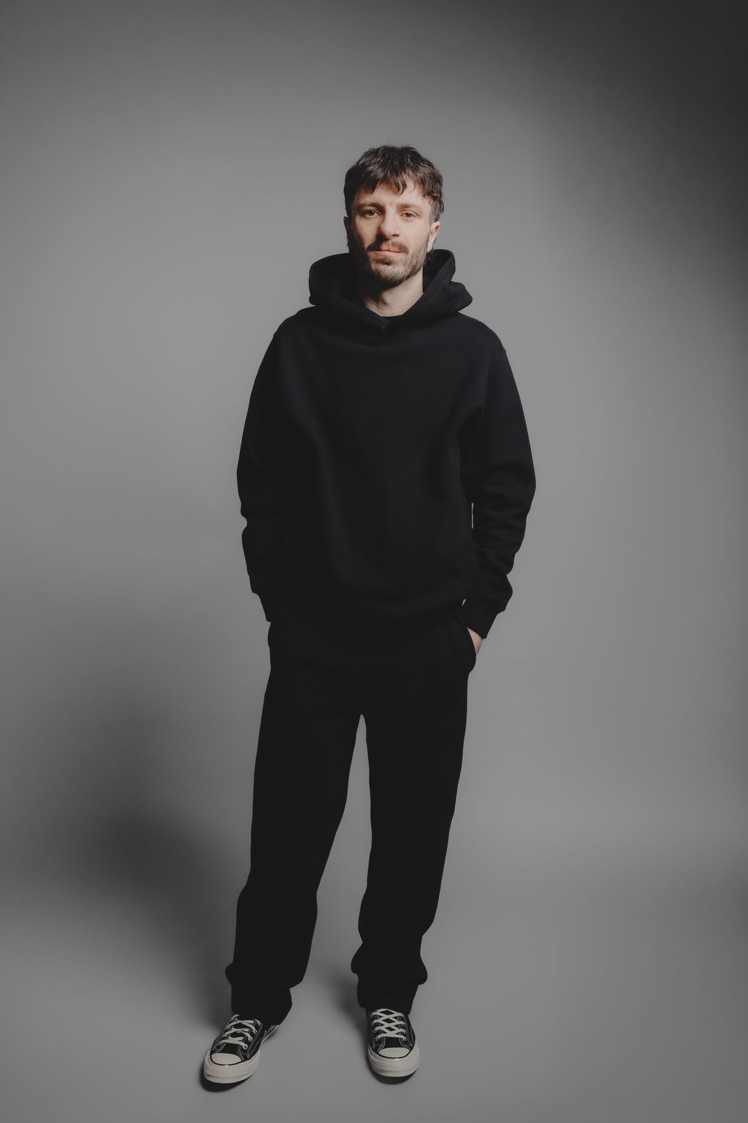 Man in black hoodie and sweatpants with Converse sneakers posing against gray background.