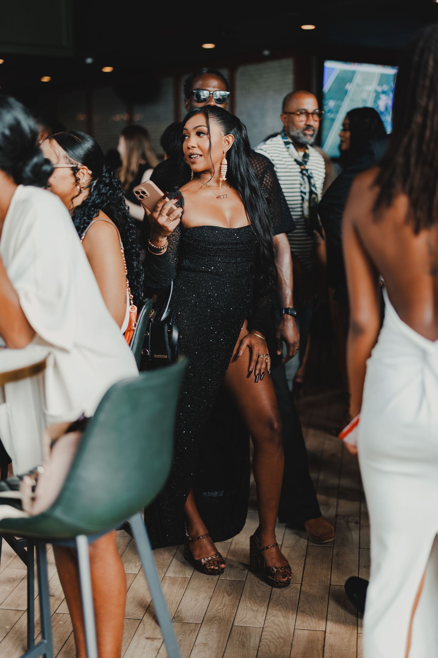 Woman in black sequin dress with thigh slit holds phone at upscale social gathering event.