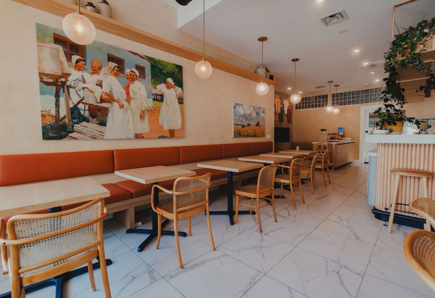 Bright cafe dining room with cane chairs, long banquette, marble floors, and chef artwork.