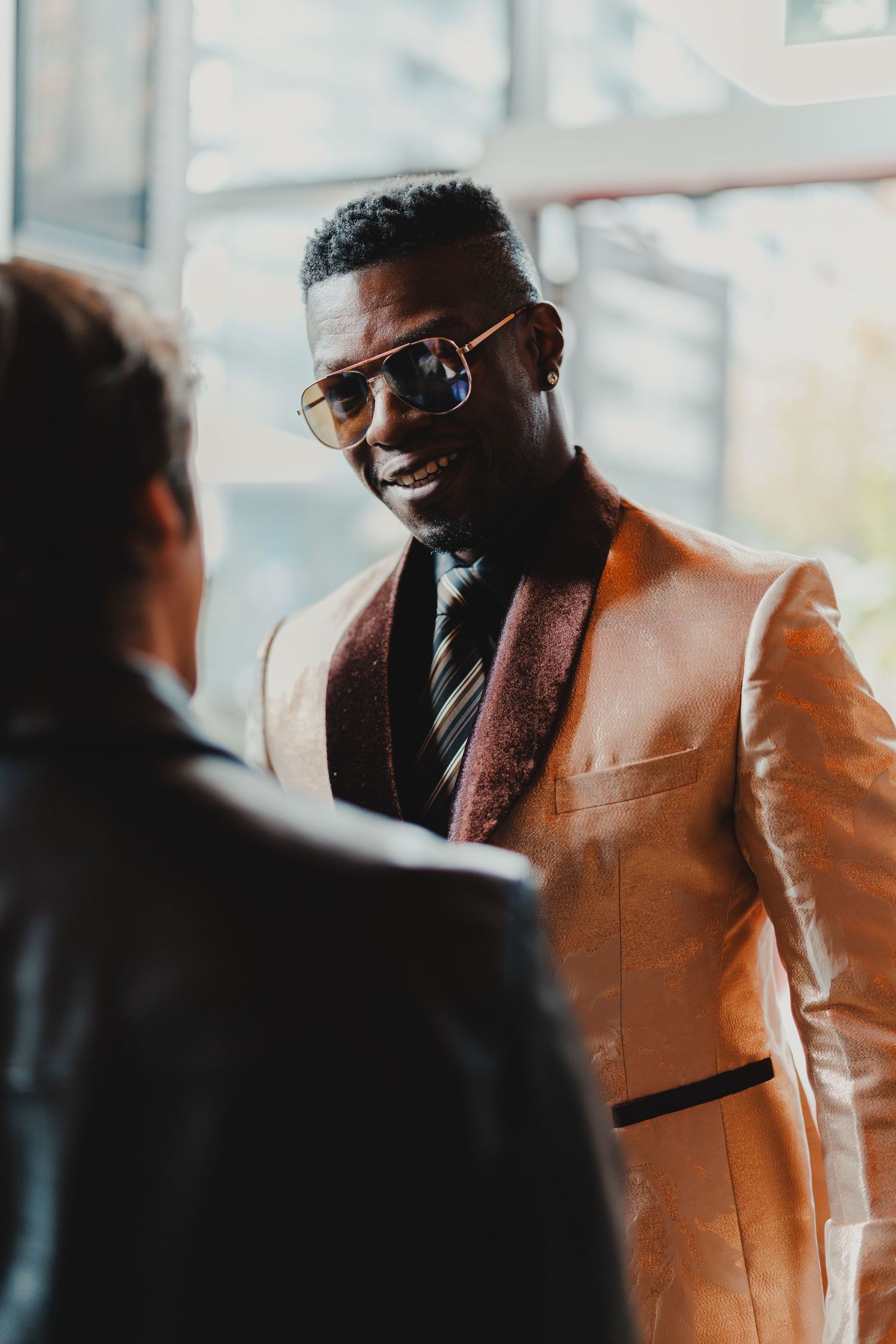 Stylish Black man in gold blazer and sunglasses smiling during a professional conversation indoors.