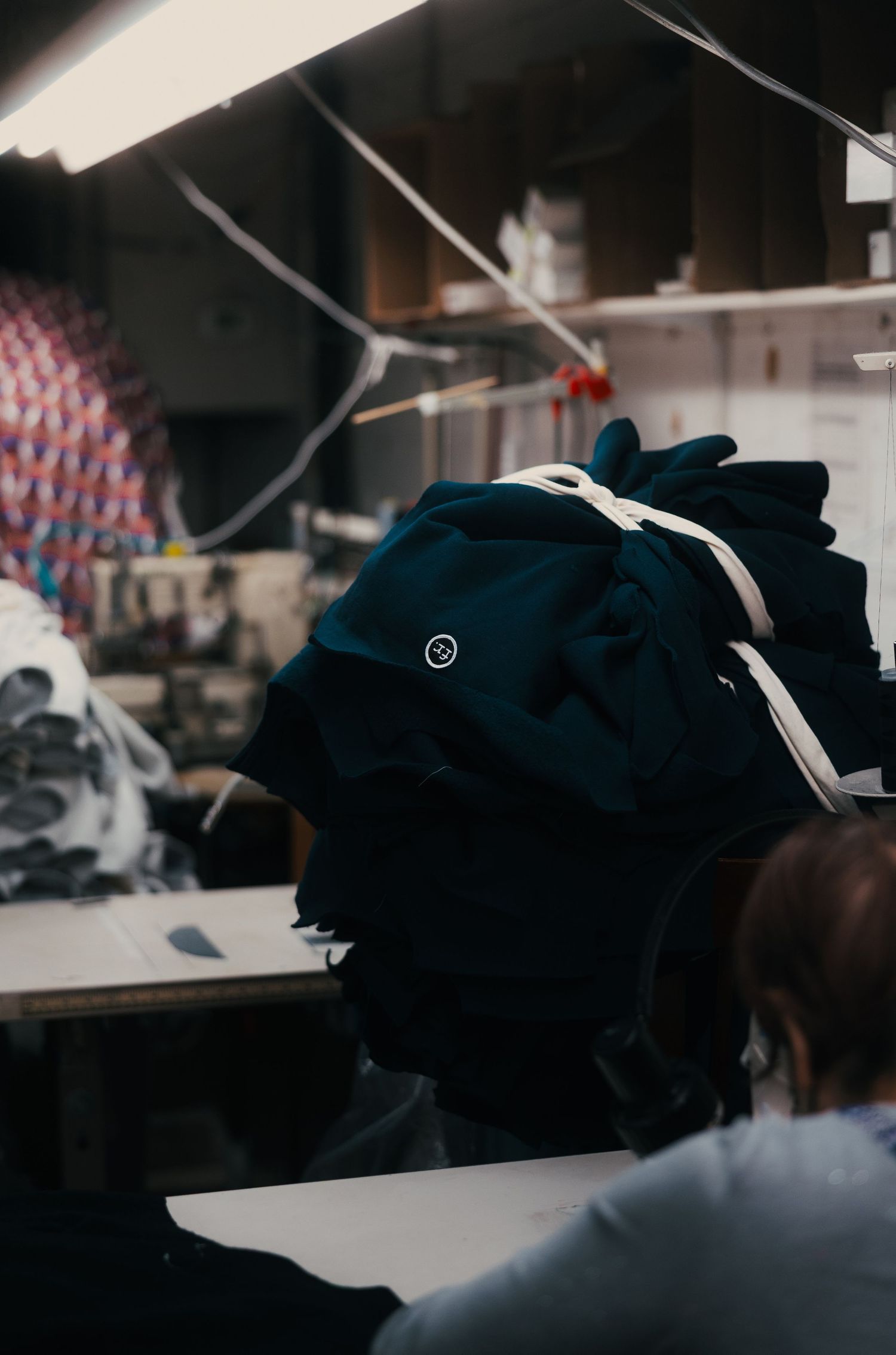 Workers in a garment factory with dark teal fabric piled high on a workstation in a dimly lit industrial sewing room.