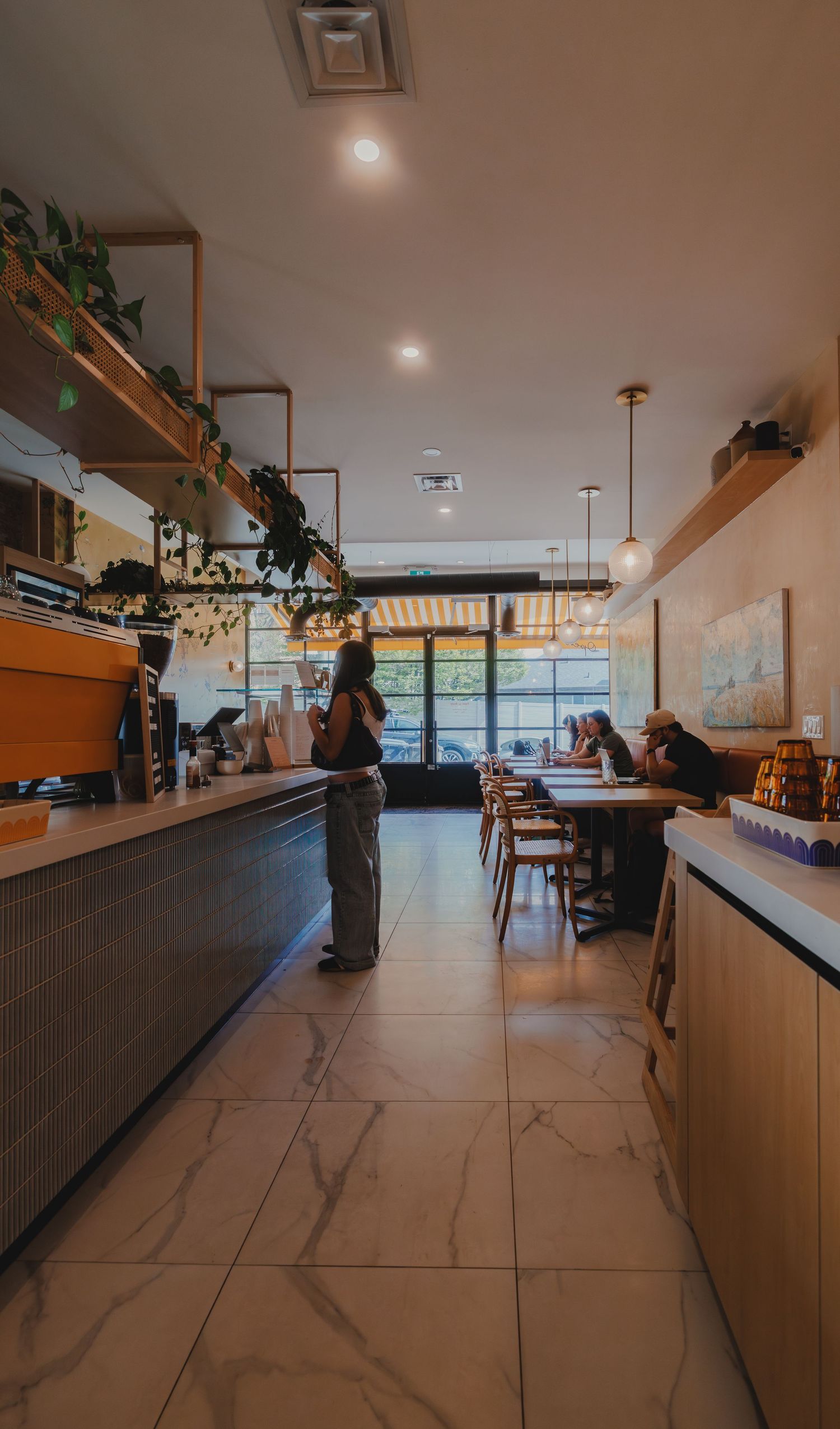 A person stands at a modern cafe counter with marble floors, hanging plants, and warm pendant lighting.