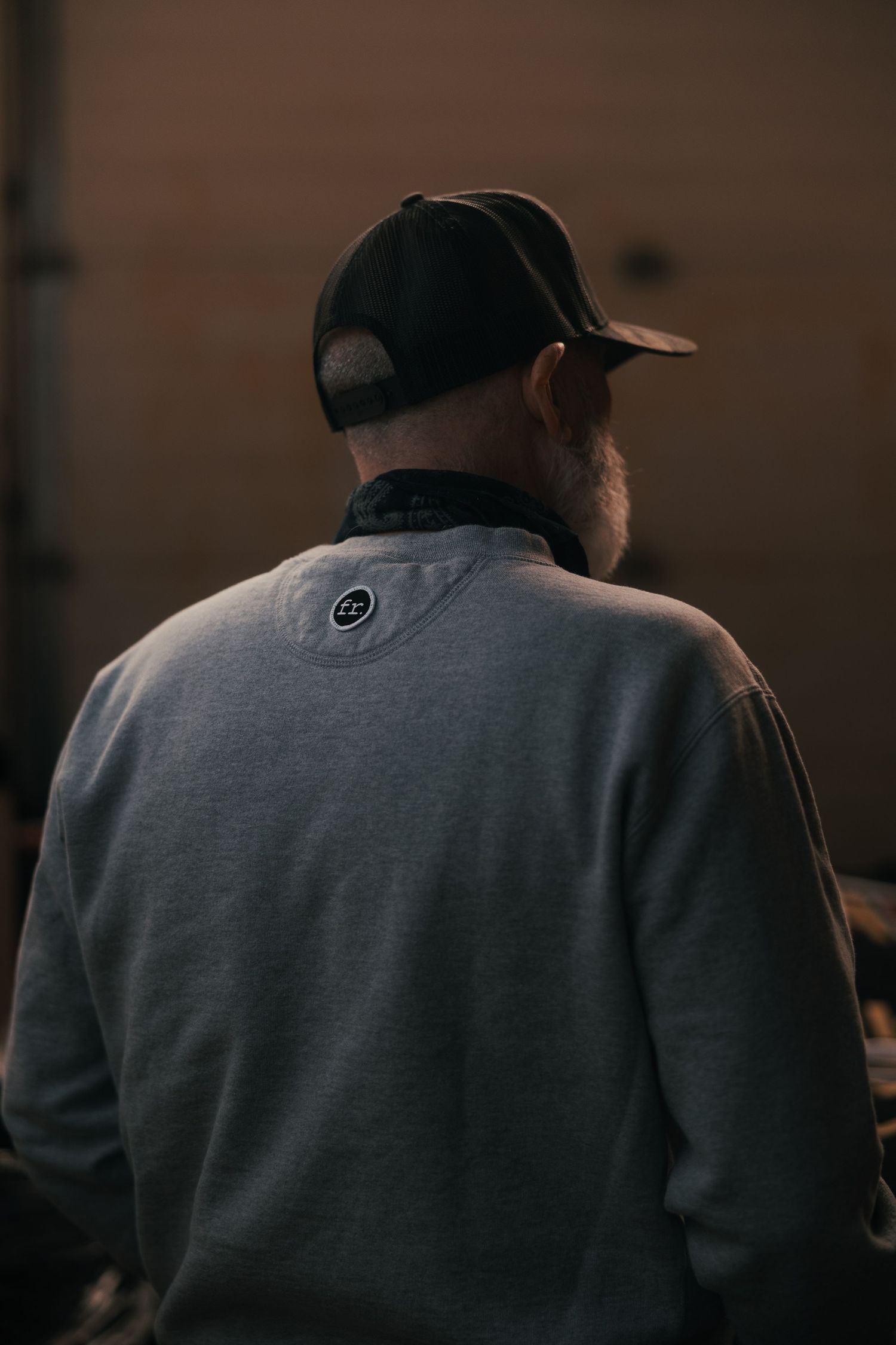 A man seen from behind wearing a gray sweatshirt and black trucker cap in a dimly lit industrial setting.