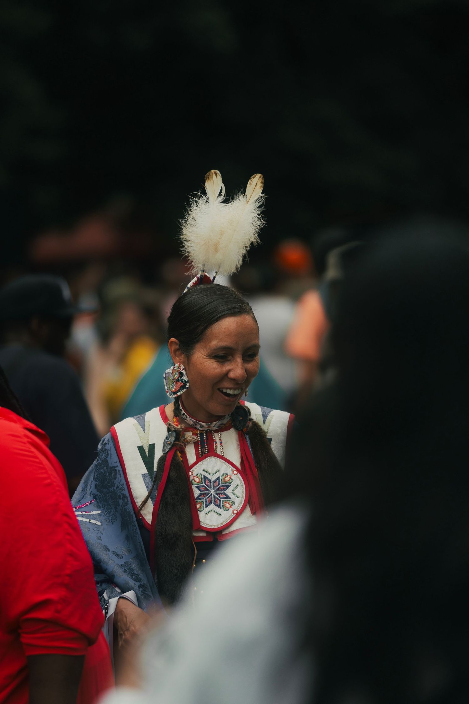 Smiling Native American woman in traditional regalia with white feather headdress at a cultural gathering.