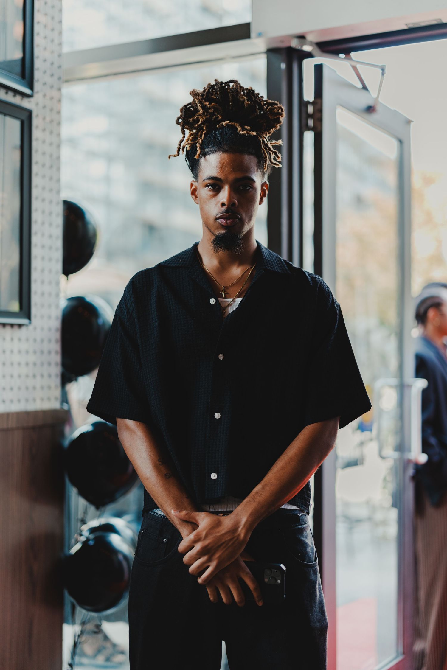 Young Black man with dreadlocks wearing a black short-sleeve button-up shirt stands in a doorway with hands clasped.