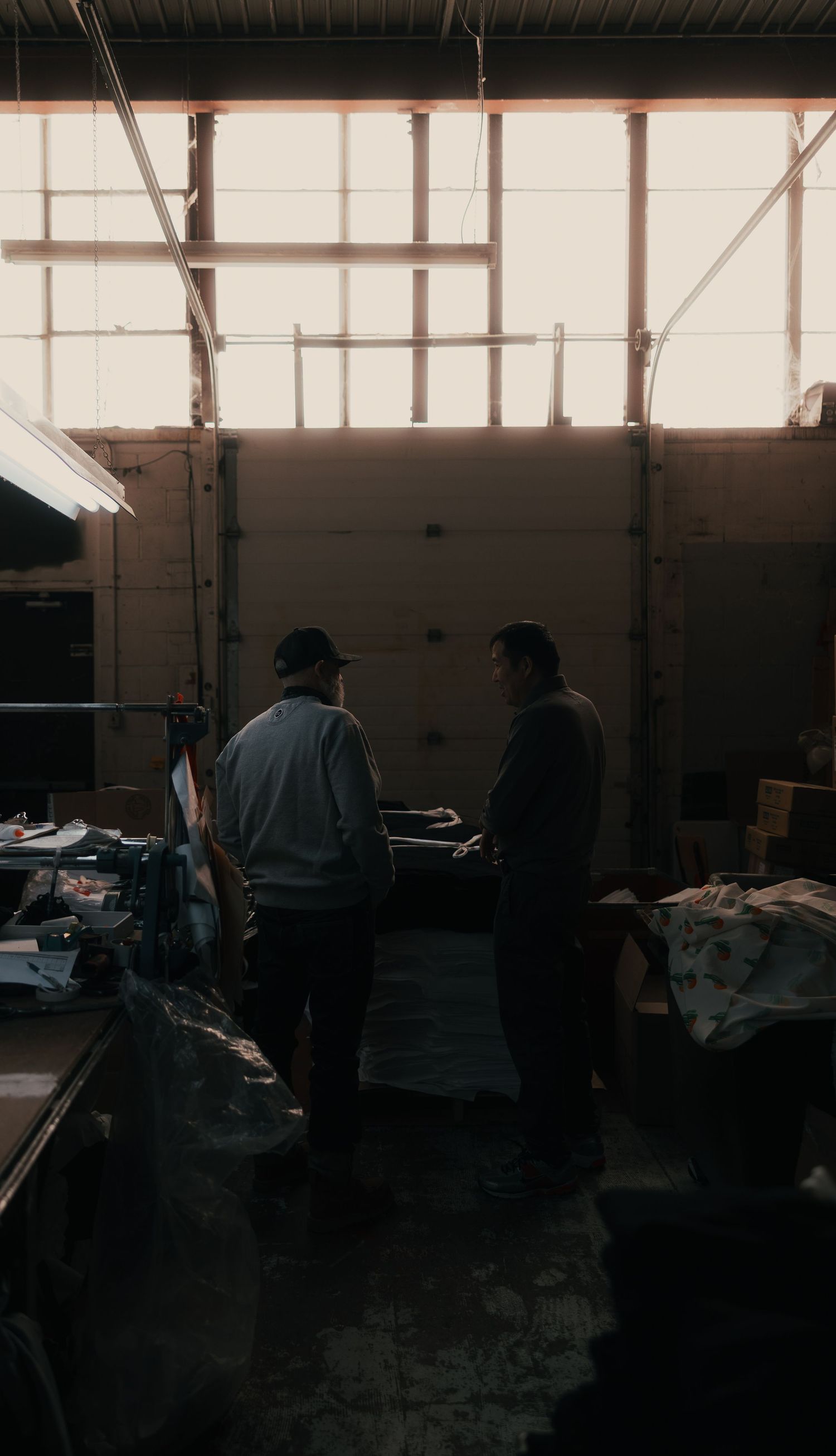 Two workers collaborating in a dimly lit industrial warehouse with large windows casting natural light from behind.