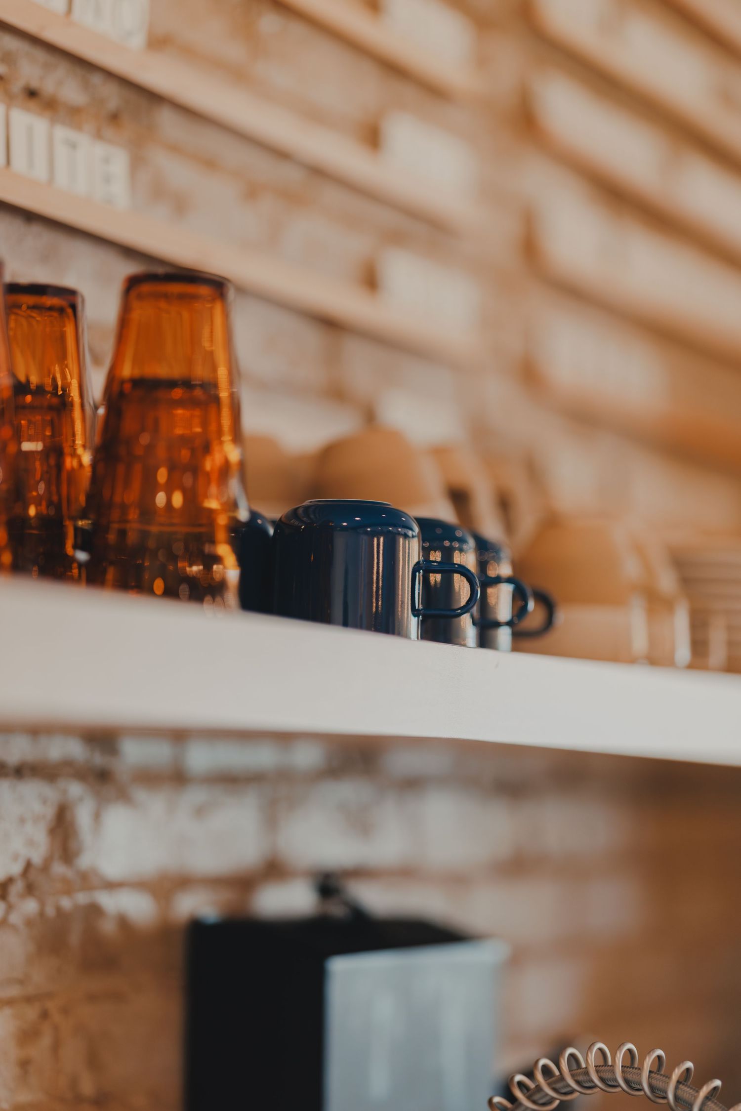 Amber glasses and dark blue mugs arranged on white shelves against a rustic brick wall backdrop.