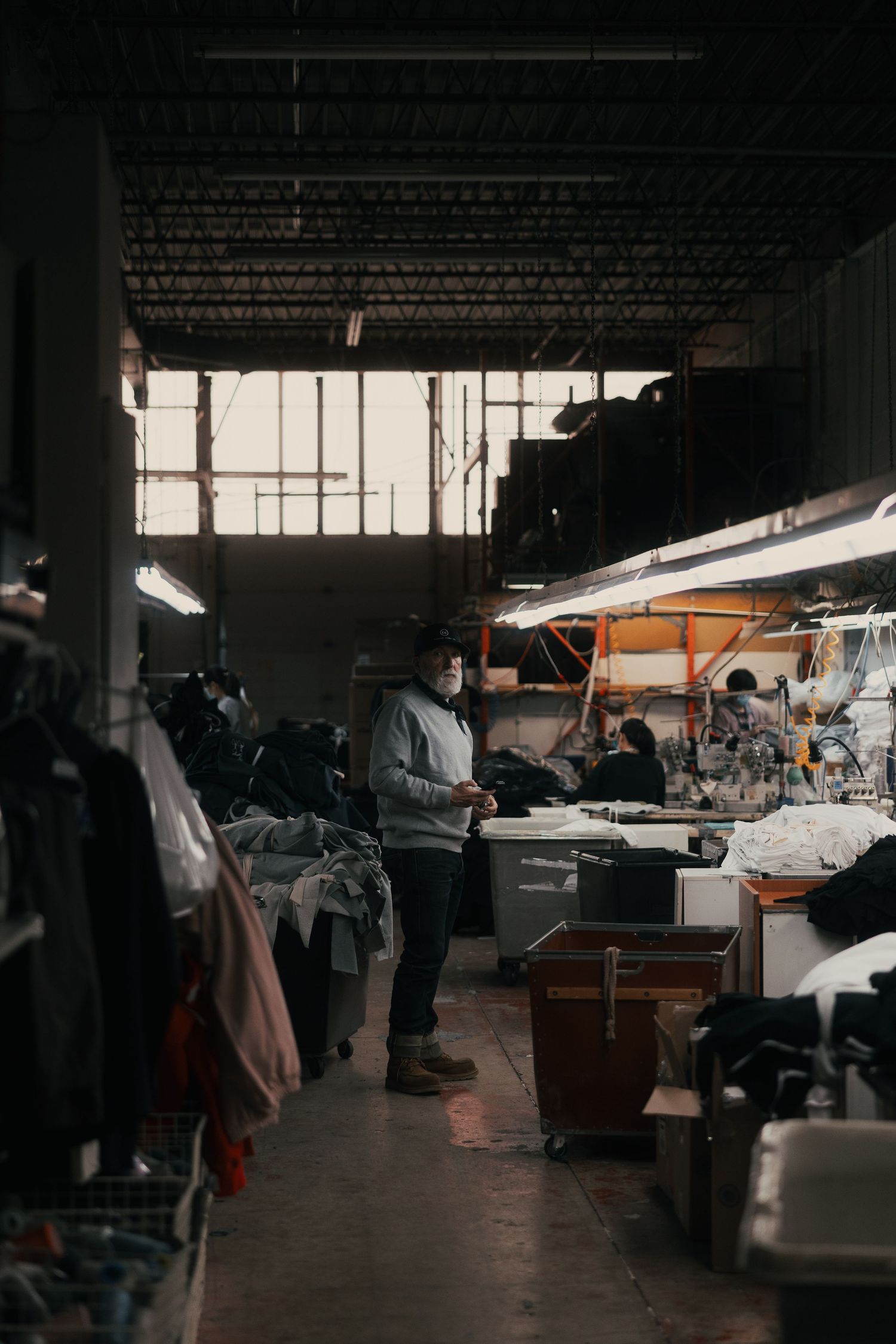 Worker in mask stands in dimly lit garment factory warehouse surrounded by clothing racks and sewing stations.