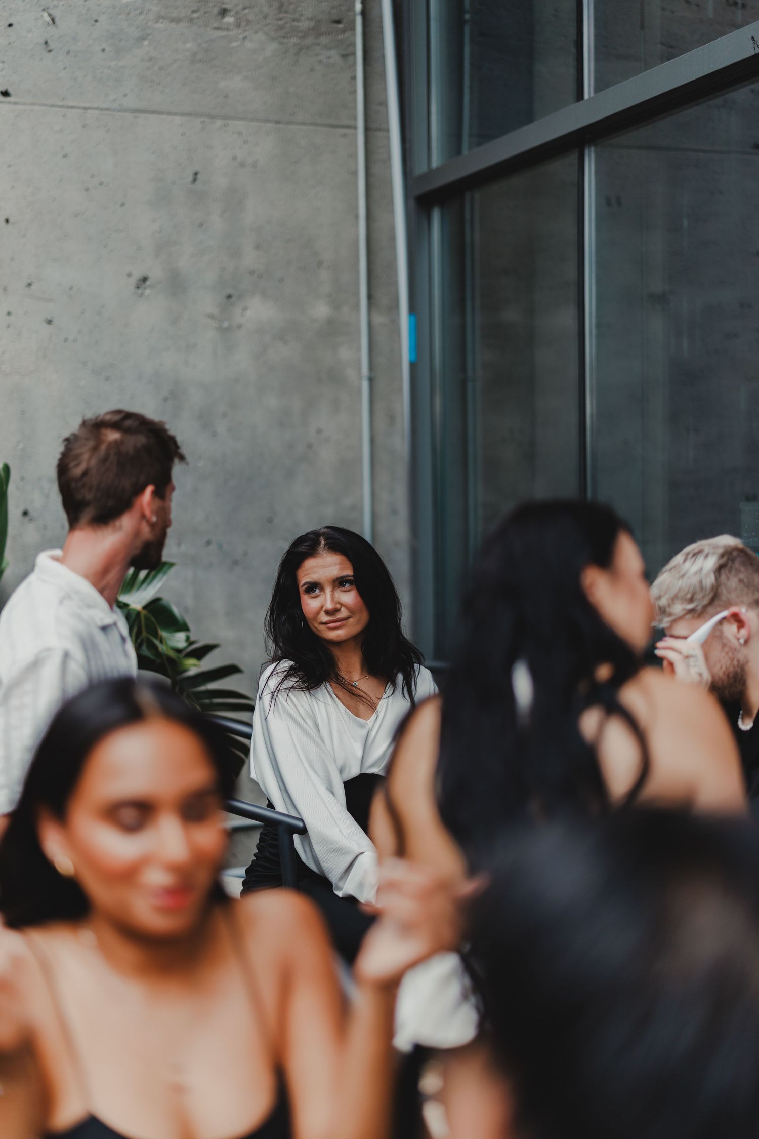 Group of young adults socializing at an indoor event, woman in white blouse smiling in focus.