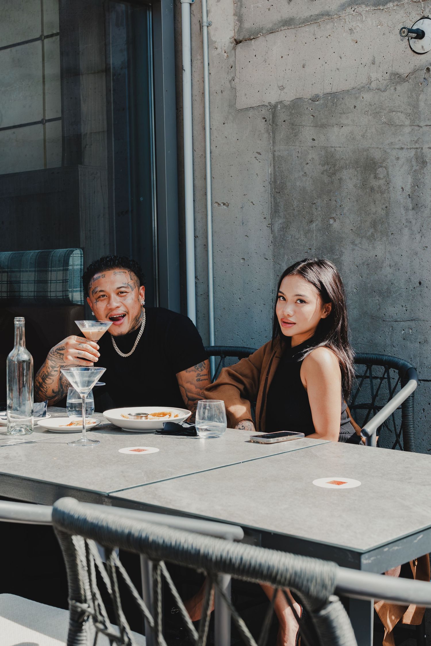 A man and woman smile at an outdoor restaurant table with martini glasses and food.