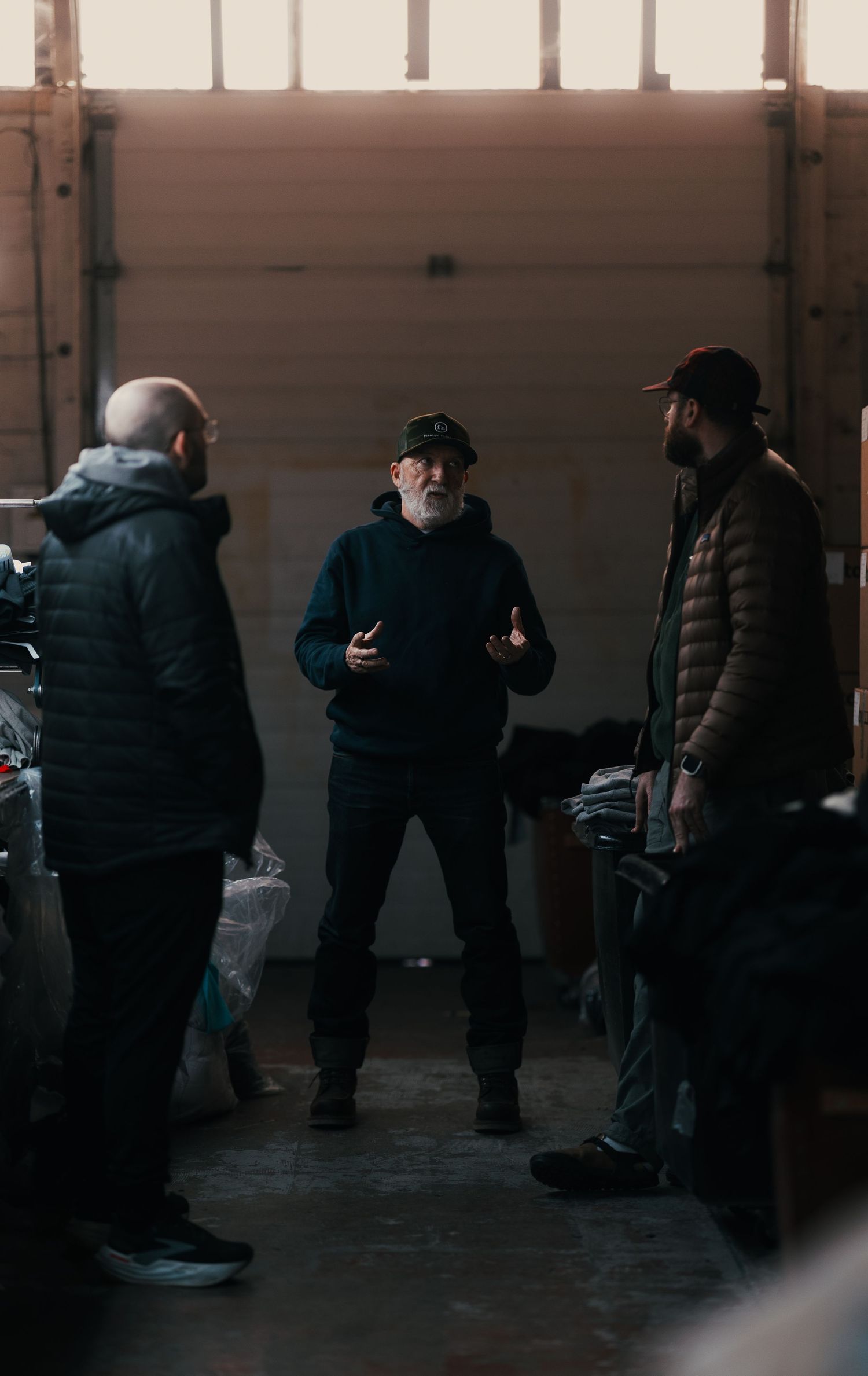 Three men in winter jackets converse inside a dimly lit warehouse, one holding a phone.