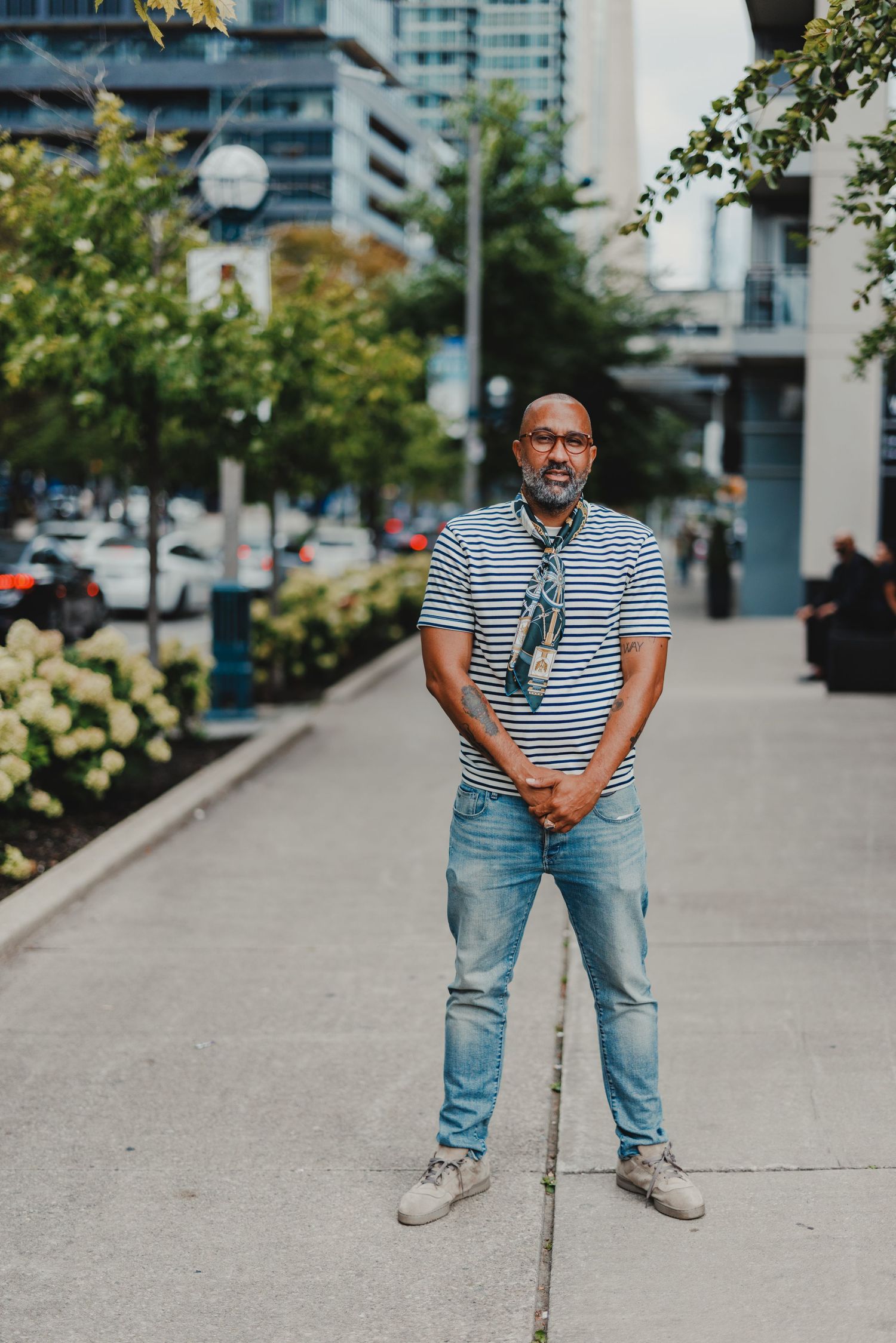 Man in striped shirt and jeans stands on urban sidewalk surrounded by city buildings and greenery.